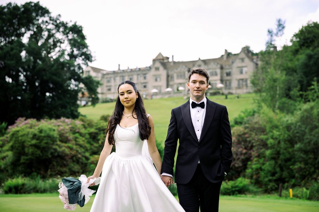 Bride in white gown and groom in black tuxedo hold hands on green lawn before a grand stone manor house.