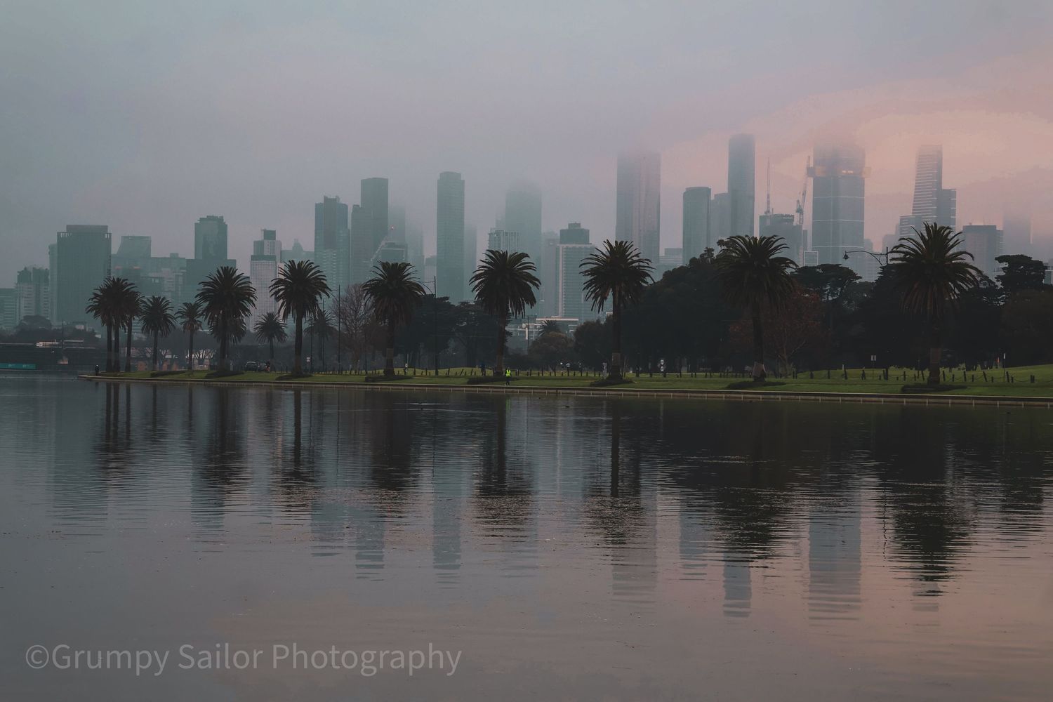 Misty Melbourne skyline reflected in calm lake waters at dusk with palm trees.