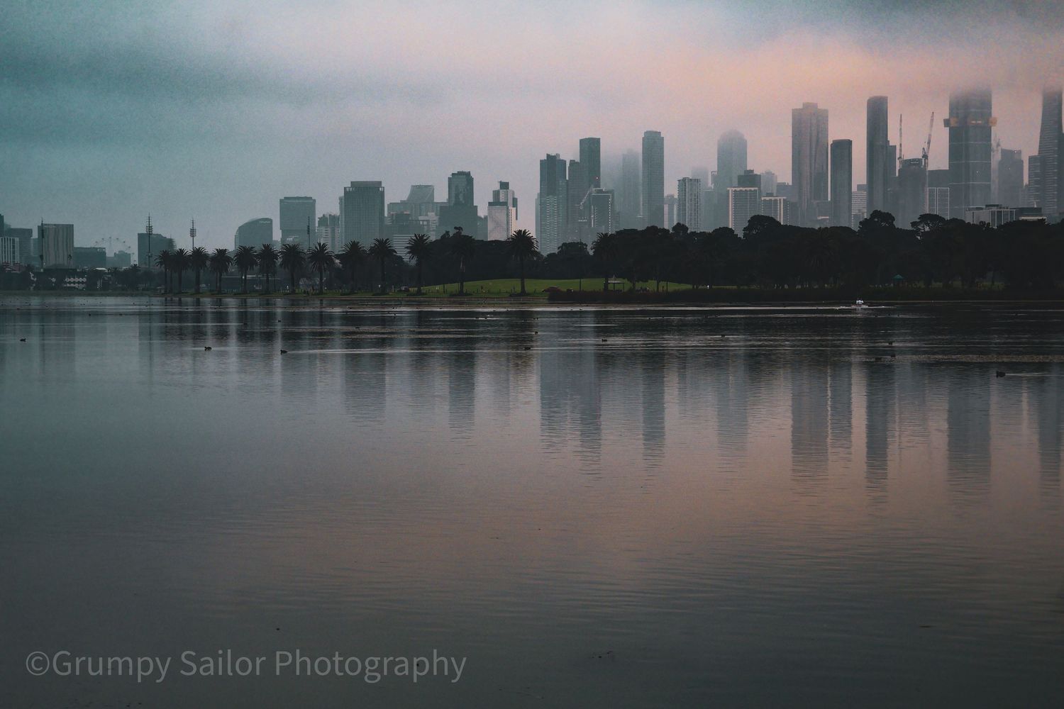 Moody Melbourne city skyline reflected in still lake waters under cloudy sky.
