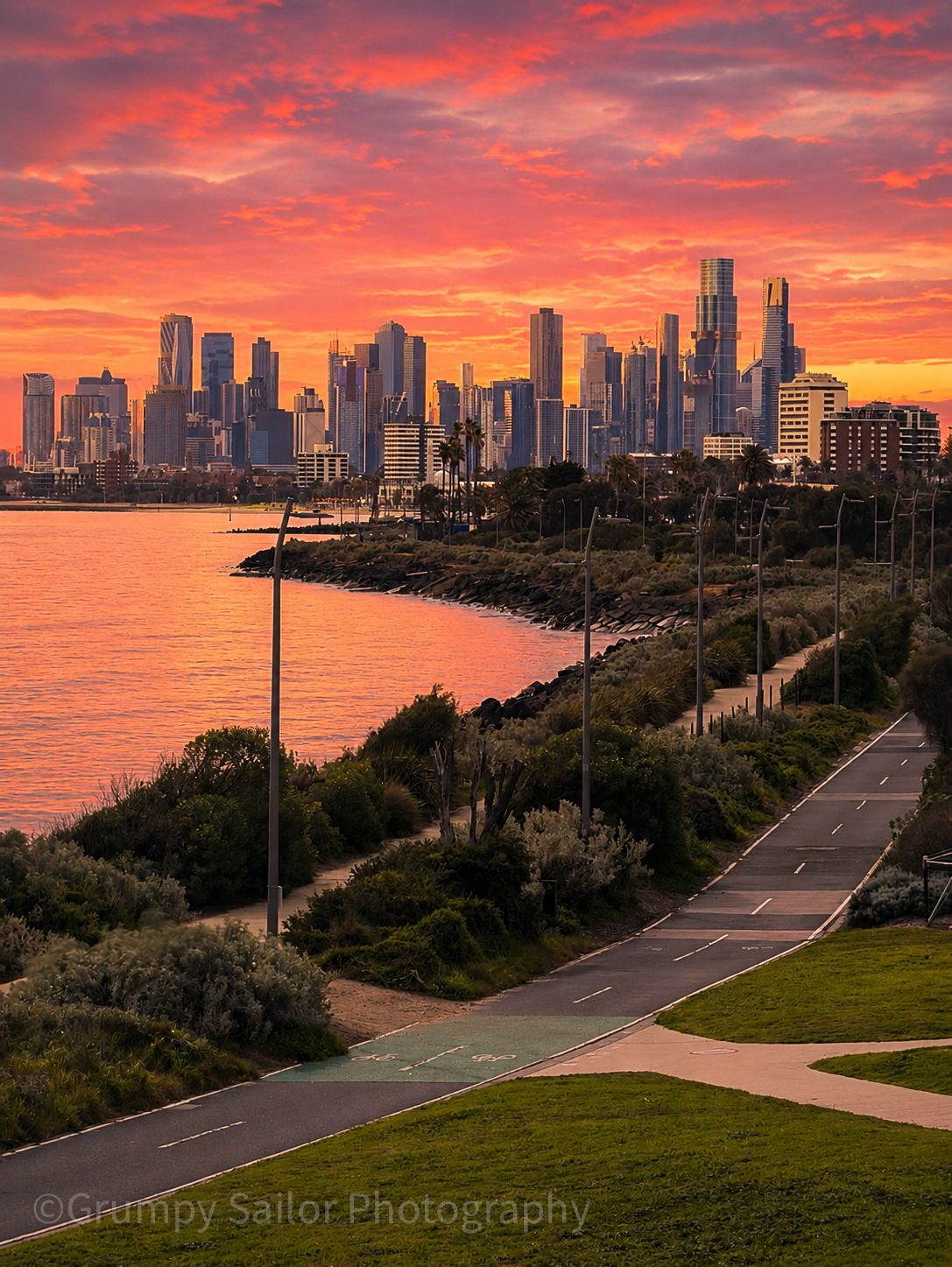 Fiery sunset over Melbourne skyline with winding coastal pathway and bay views.