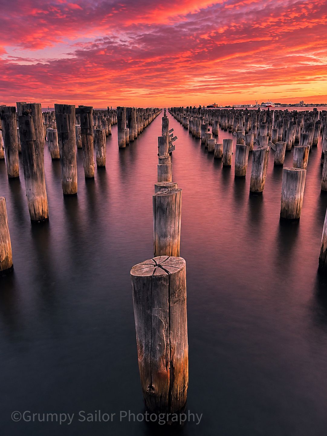 Symmetrical rows of weathered wooden pylons extending into calm bay under vivid red sunset.