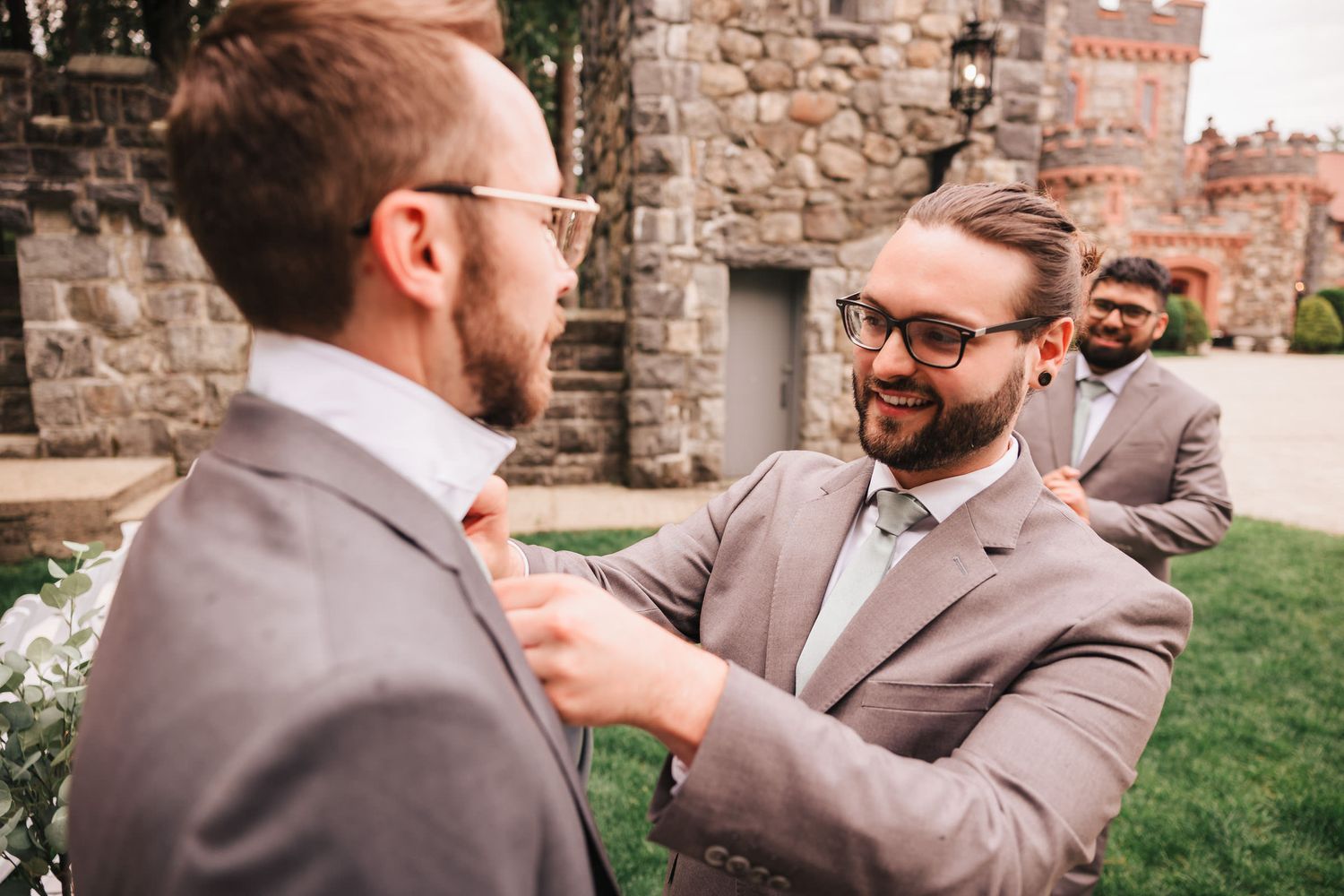 A groomsman adjusting another's collar and smiling, with the stone towers of Searles Castle in the background.
