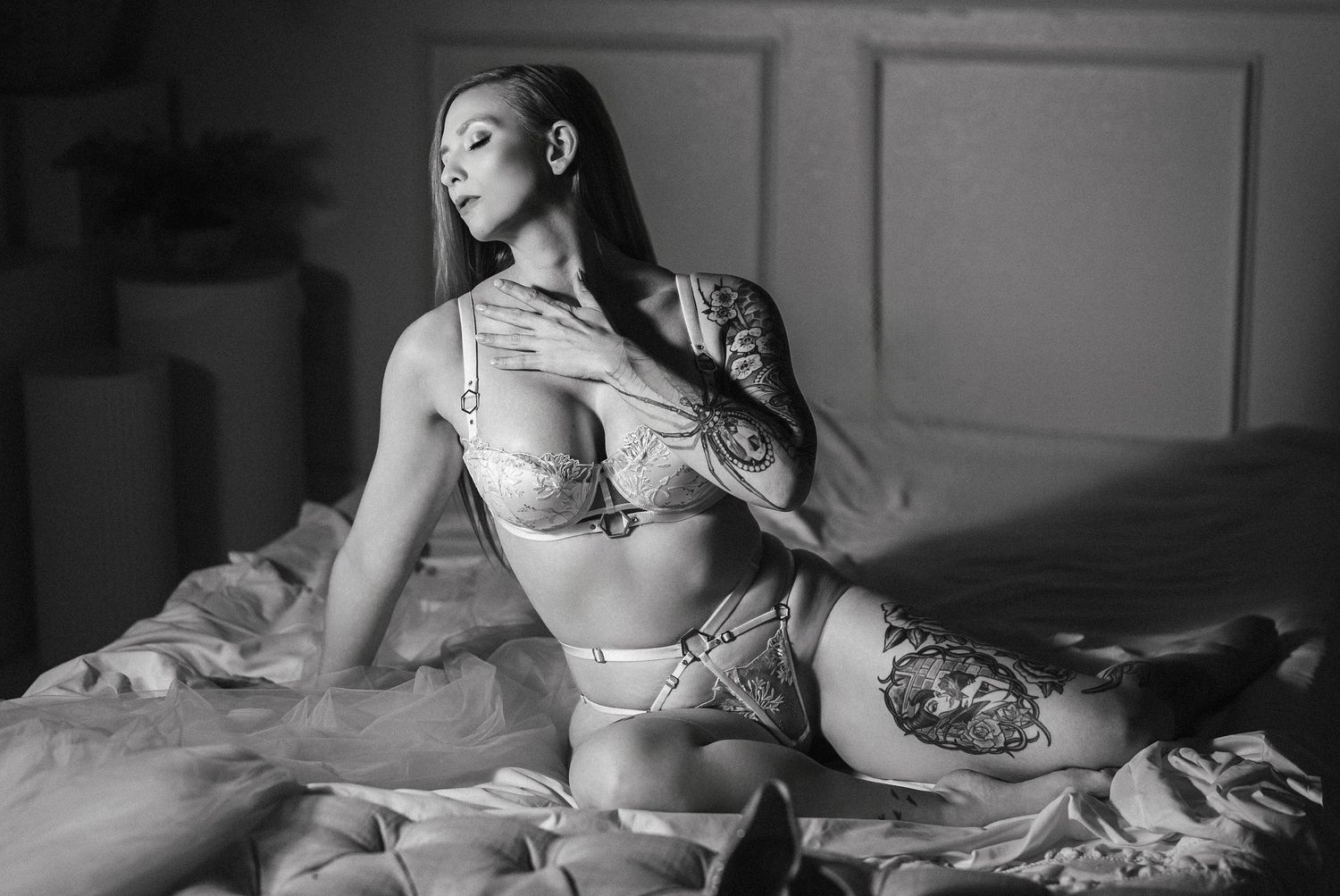 Black and white boudoir photo of a tattooed woman posing gracefully on a bed in elegant lingerie.