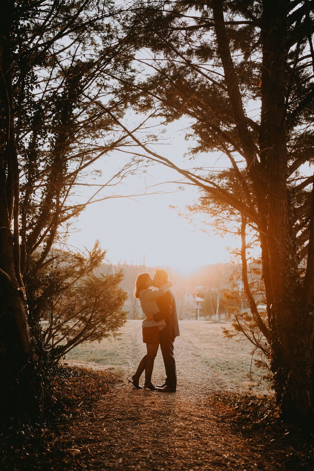 Couple silhouetted against golden sunset light, embracing on a forest path surrounded by tall trees in autumn.