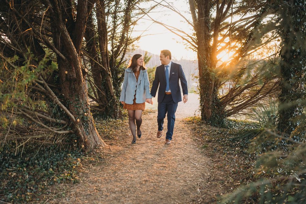 Couple holding hands walking on a sunlit forest path surrounded by tall trees at golden hour.