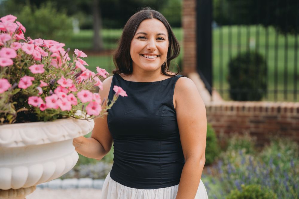 Smiling young woman in black top and white skirt standing beside a stone planter with pink petunias outdoors.