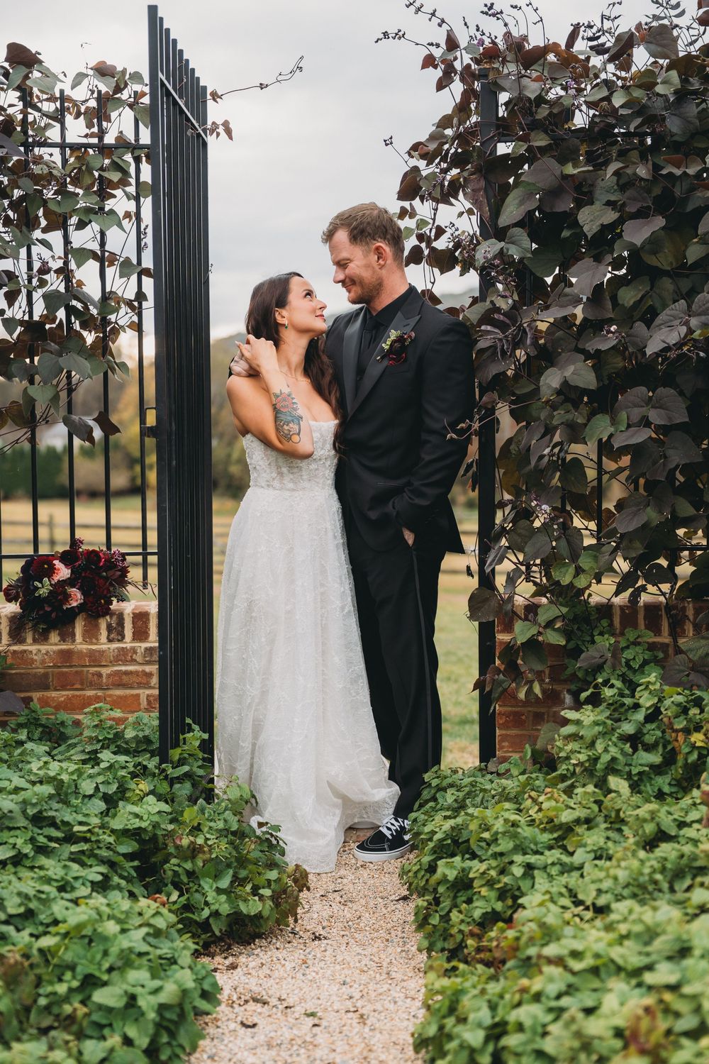 Bride and groom share a romantic moment in a garden gate, surrounded by lush greenery, wearing wedding attire.