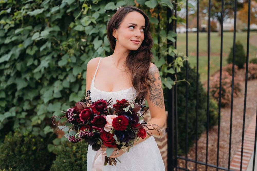 Bride in white gown holding dark red and burgundy wedding bouquet standing near iron gate with ivy backdrop.