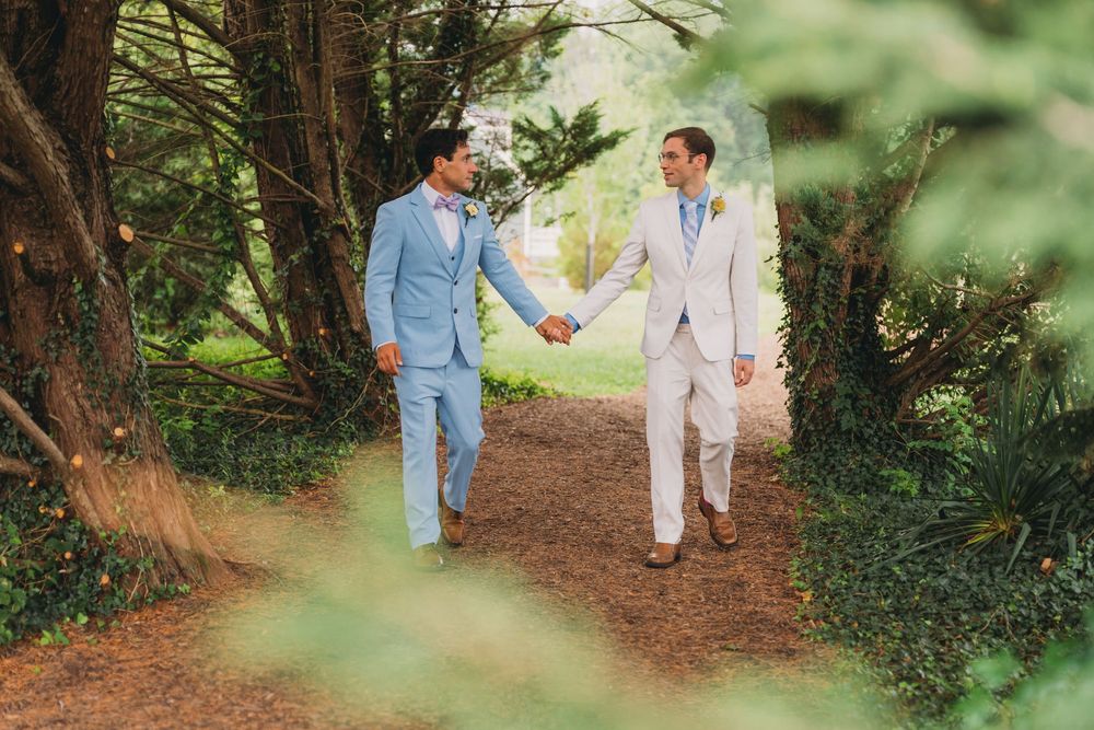 Two grooms in light blue and white suits hold hands while walking along a tree-lined path at their outdoor wedding.