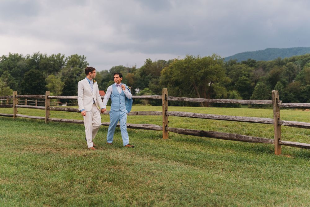 Two grooms walk hand-in-hand along a wooden fence on a green farm with mountain views on their wedding day.