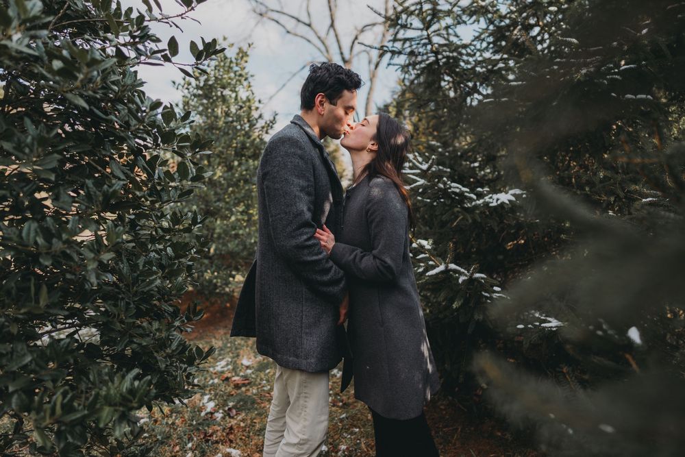 Couple sharing a romantic kiss surrounded by lush green trees in a winter outdoor setting.