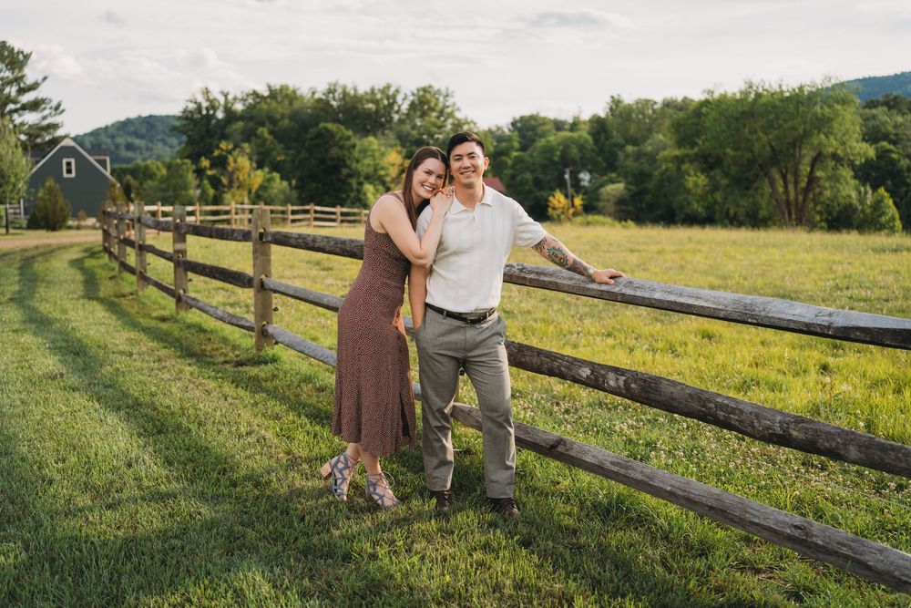 Couple embracing by a rustic wooden fence in a green countryside farm setting on a sunny day.