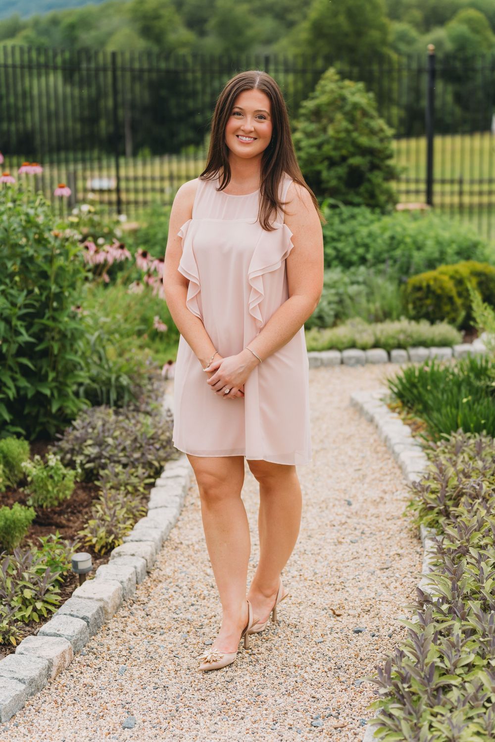 Young woman in pink ruffle dress posing on garden path surrounded by lush greenery and flowers.