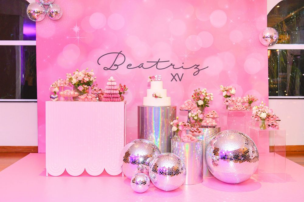 Pink quinceañera dessert table for Beatriz XV with disco balls, floral arrangements, and a white cake on metallic pedestals.