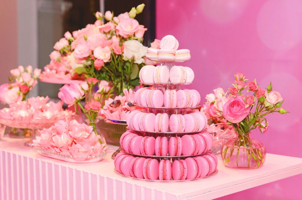 Pink macaron tower display with floral arrangements at a dessert table celebration.
