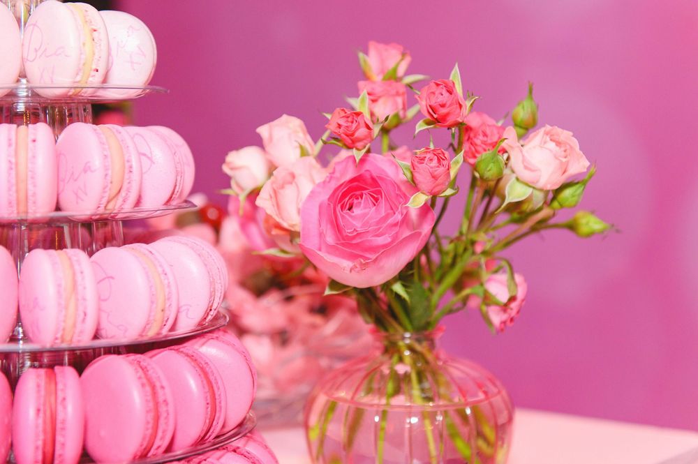 Pink macarons on tiered stand beside a glass vase with pink roses and flowers against a pink background.