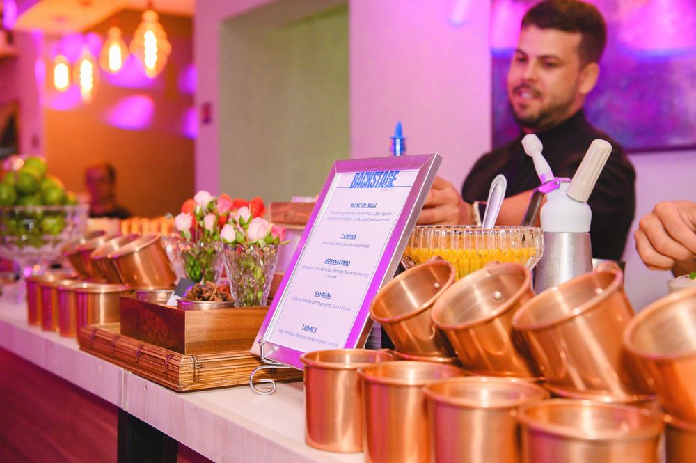 Bartender stands behind a bar lined with copper Moscow mule mugs, a menu sign, and floral arrangements at an event.