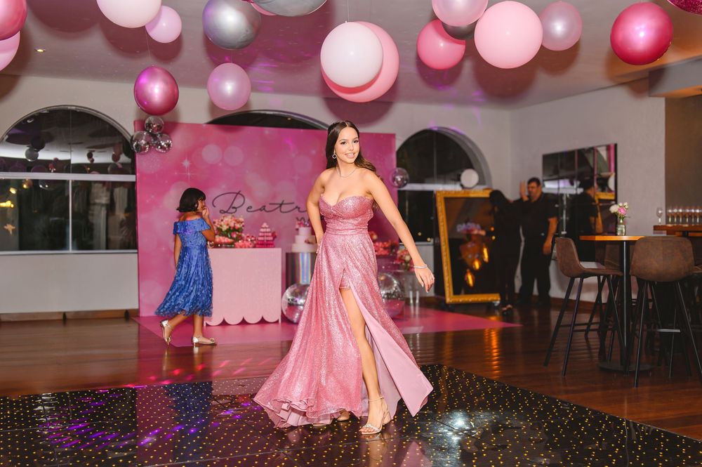 Young woman in pink satin gown dancing at elegant quinceañera party with pink balloon decorations.