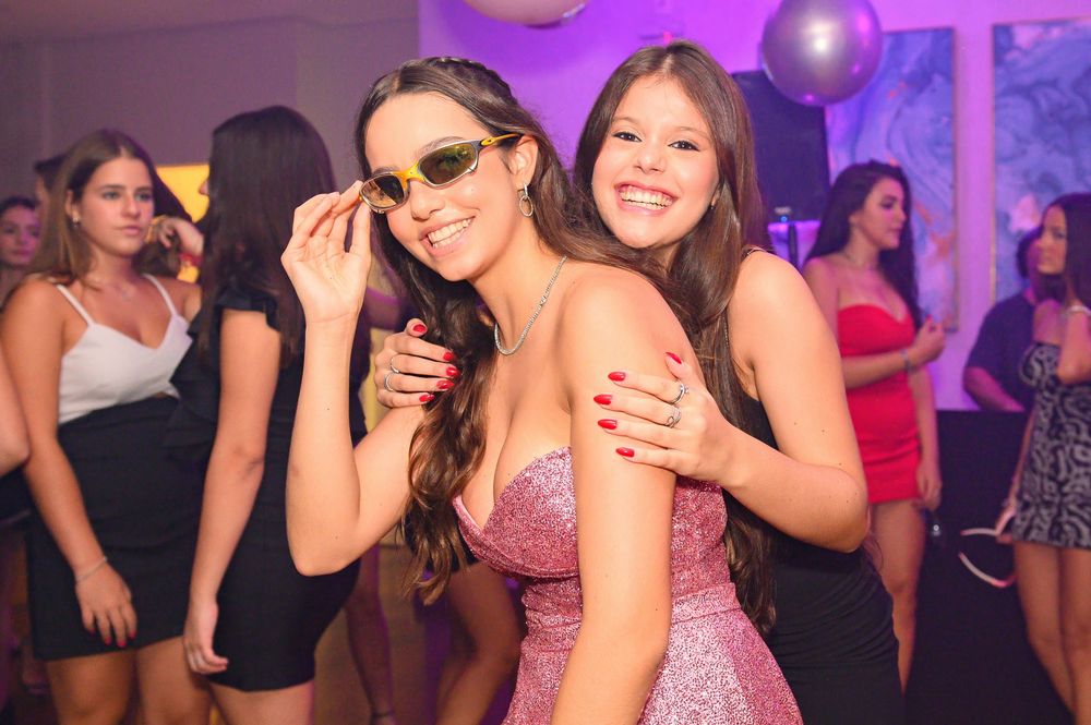 Two young women posing together at a party, one wearing yellow sunglasses and a pink sparkly dress.