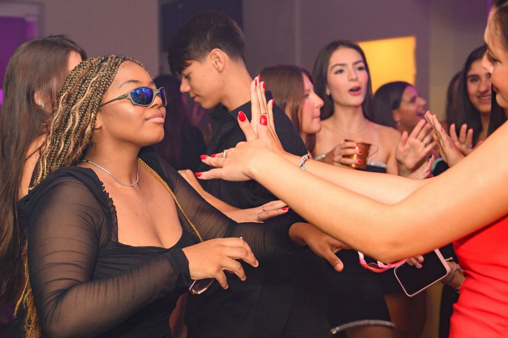 People dancing and socializing at a lively party, with a woman in black mesh top and sunglasses in the foreground.