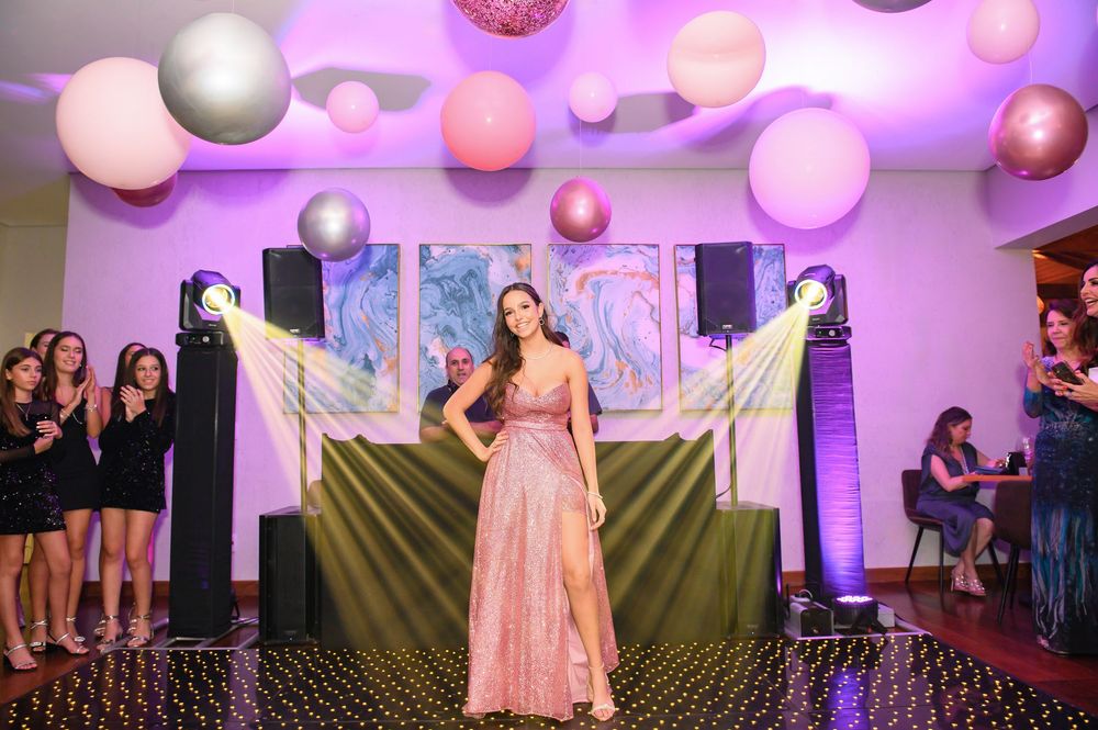 A woman in a pink gown poses on a dance floor at an elegant party with large balloons and purple lighting.