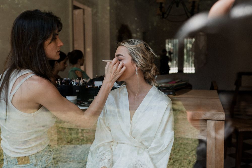 Makeup artist applying eye makeup to a bride in a white robe during wedding preparations indoors.