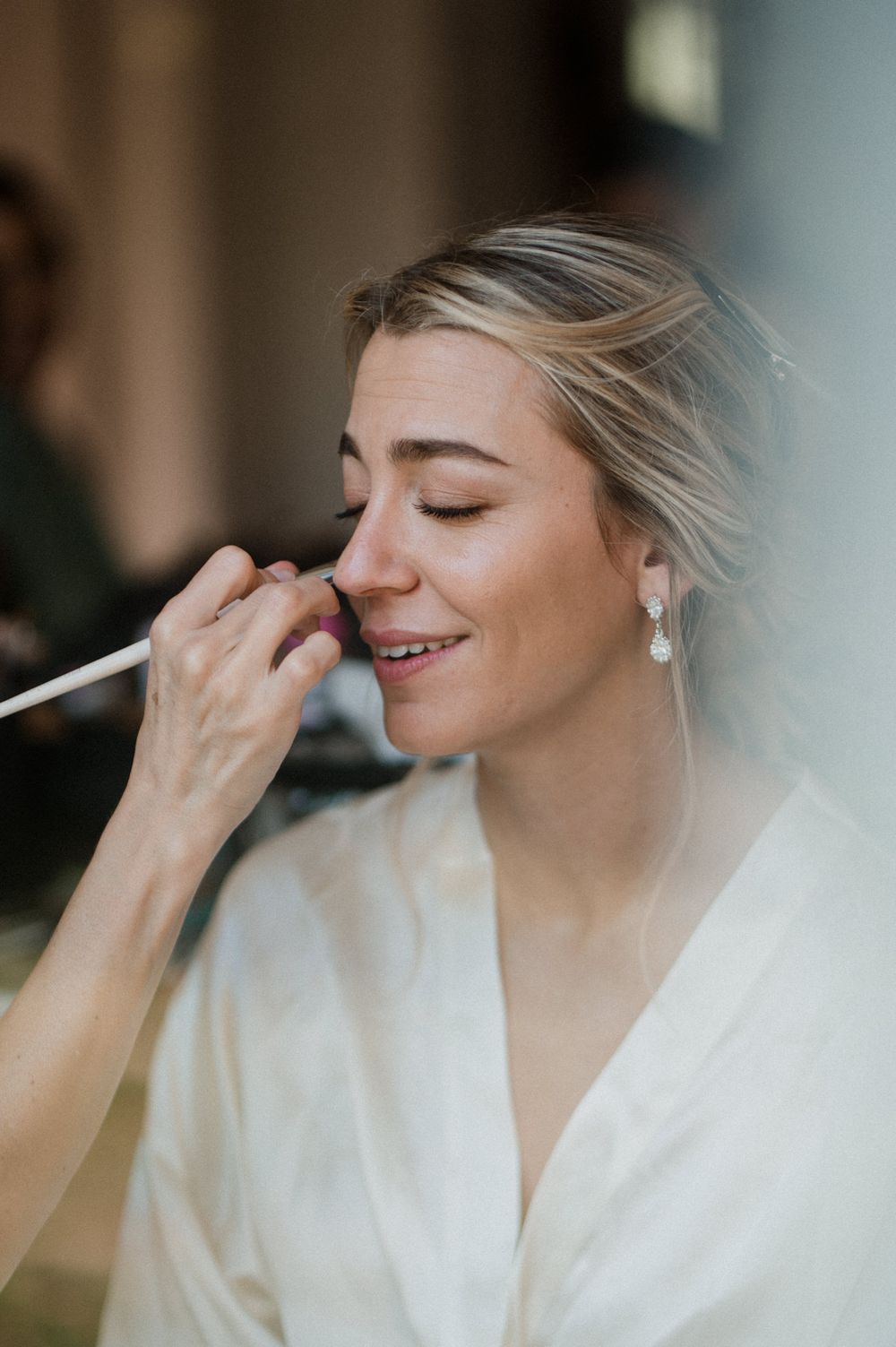 Bride smiling with eyes closed while makeup artist applies foundation with a brush during wedding preparations.