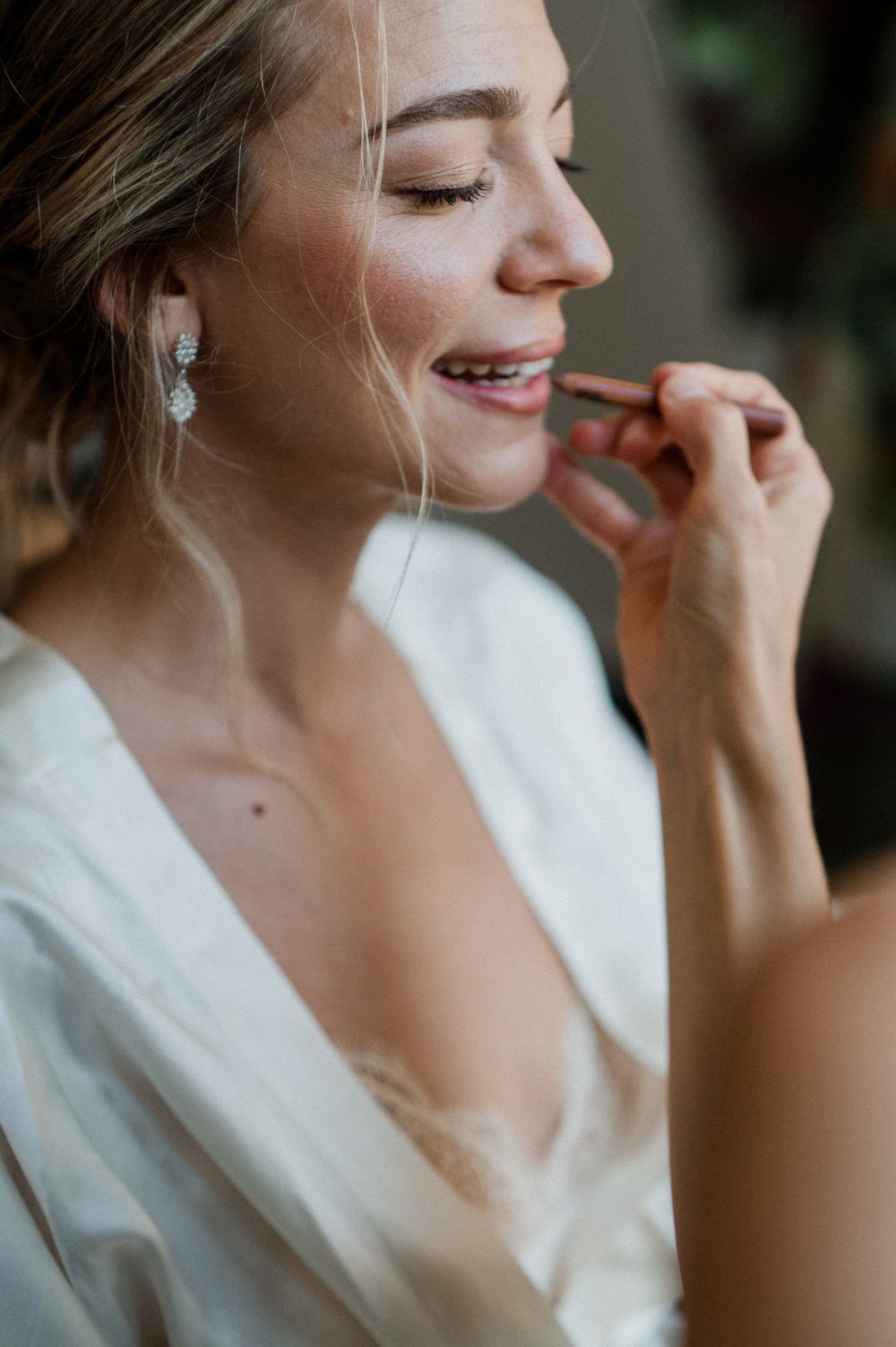 Smiling bride applying lipstick while getting ready, wearing elegant drop earrings and a white satin robe.