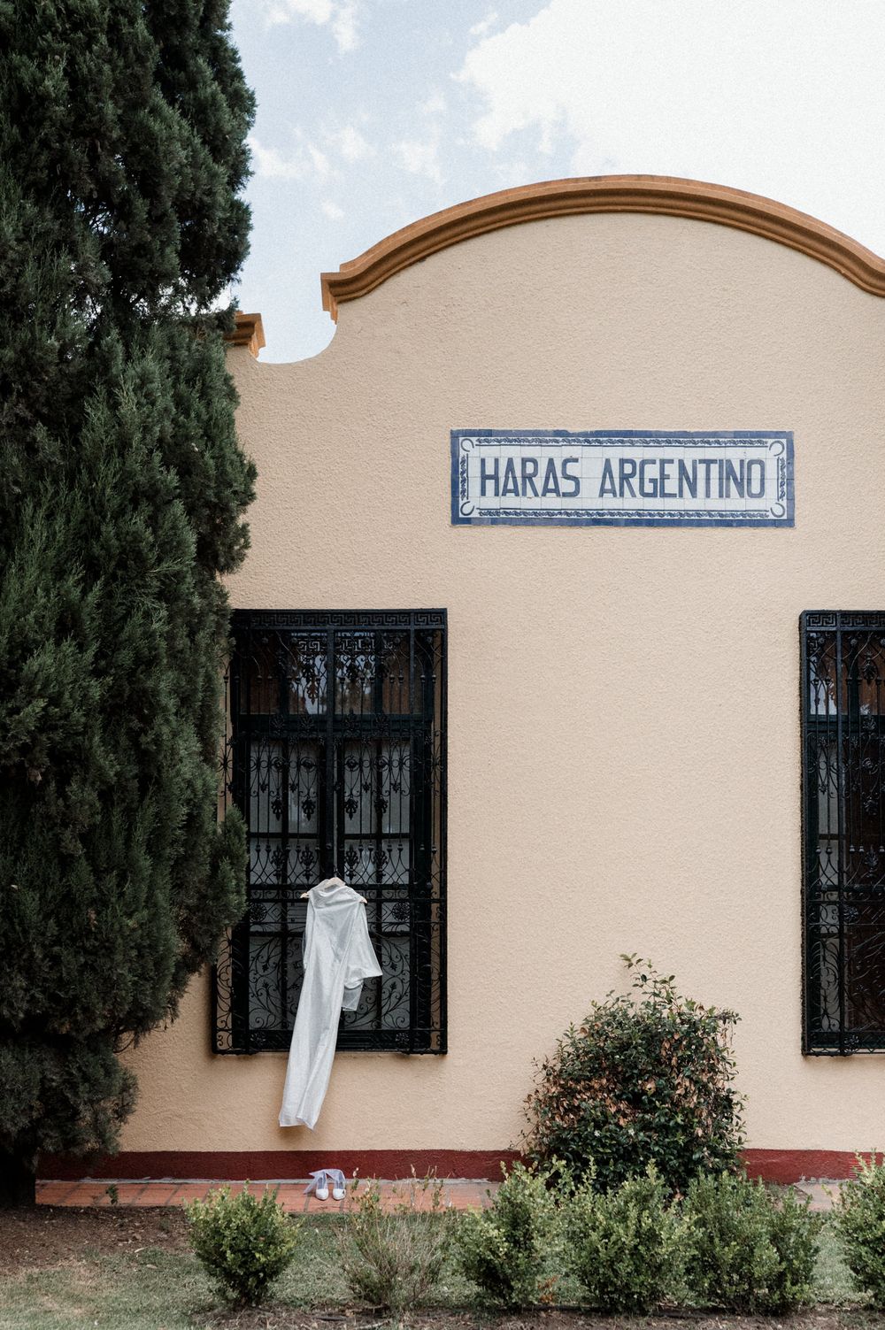 White wedding dress hanging on iron-barred window of Haras Argentino building facade with cypress trees.