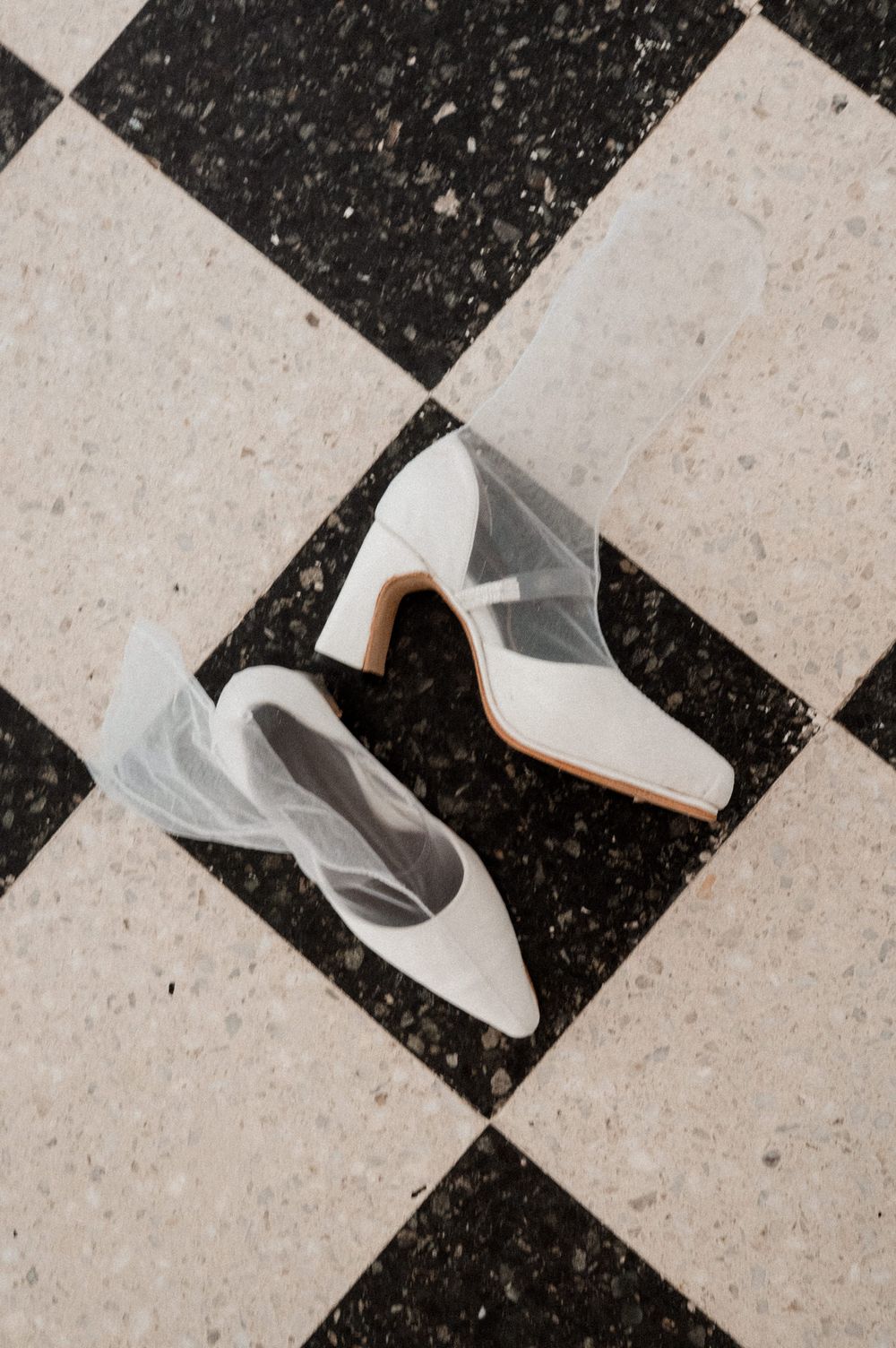 White bridal heels with sheer tulle bow detail on black and white checkered tile floor.