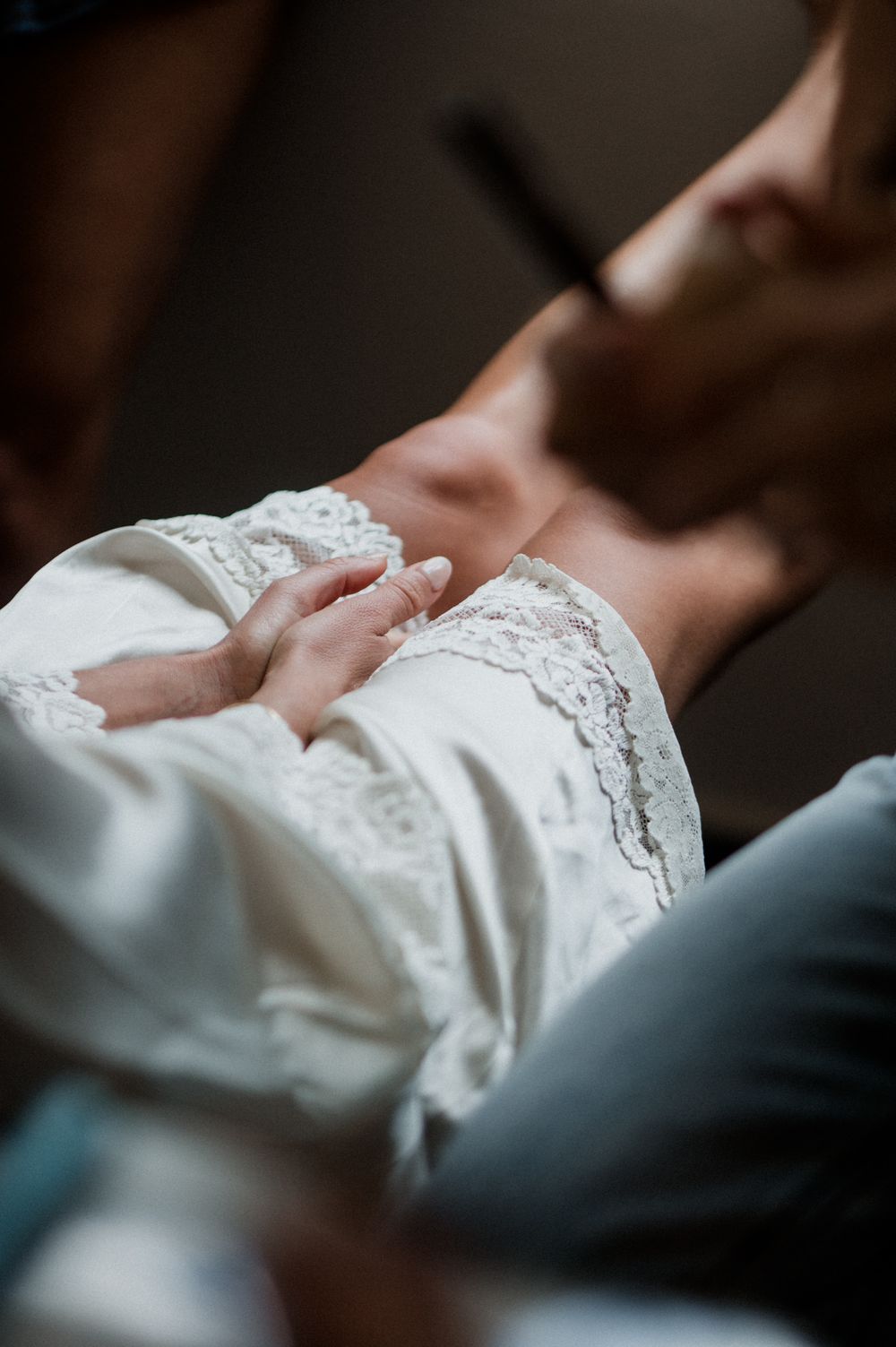 Close-up of a bride in white lace wedding dress with groom's hands gently holding her waist.