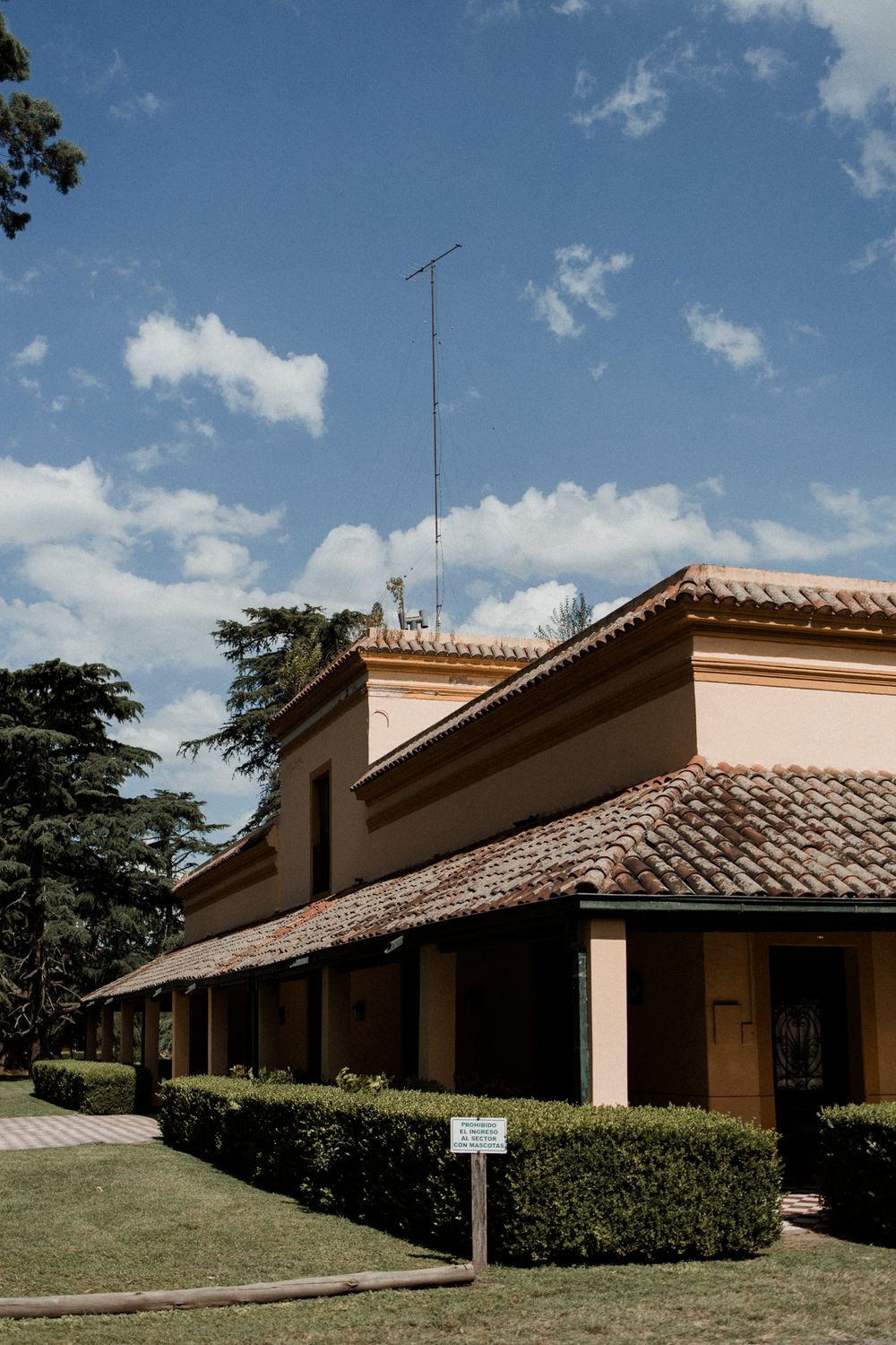 Yellow colonial building with terracotta tile roof, trimmed hedges, and TV antenna against a partly cloudy blue sky.