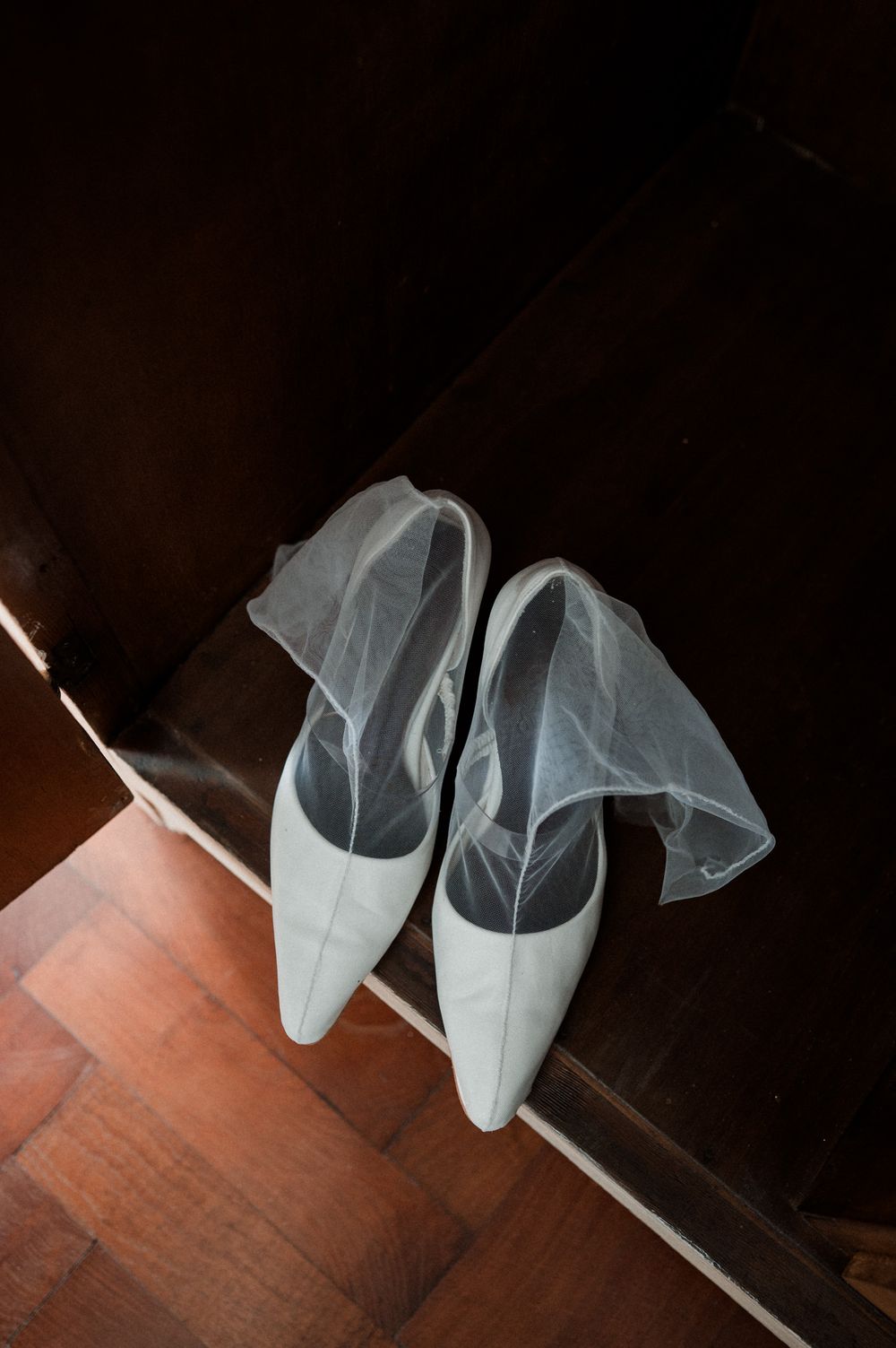 White bridal heels with delicate tulle veil detail resting on wooden parquet floor near dark doorway.