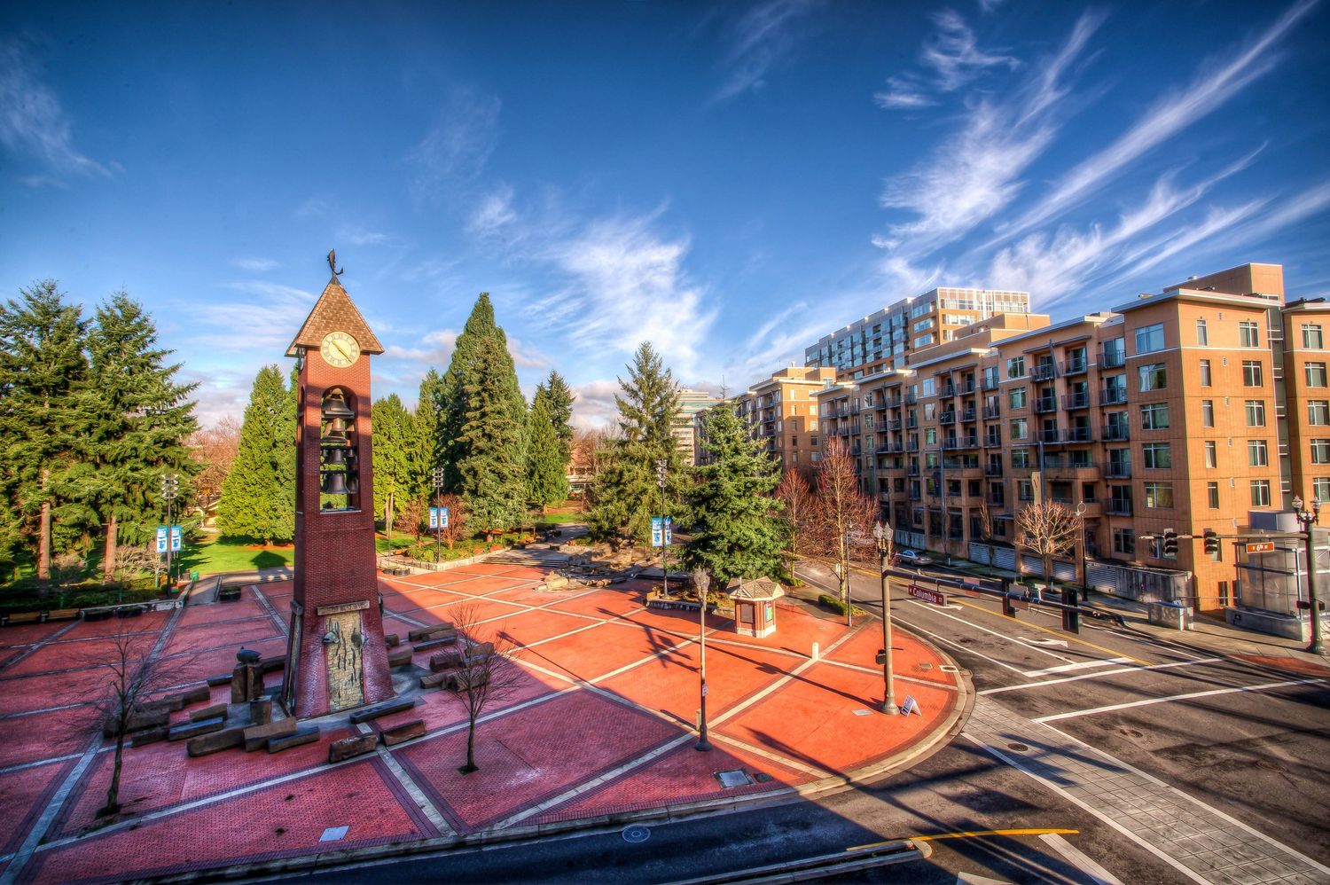 Clock tower in a red brick plaza surrounded by evergreen trees with modern apartment buildings and blue sky in background.