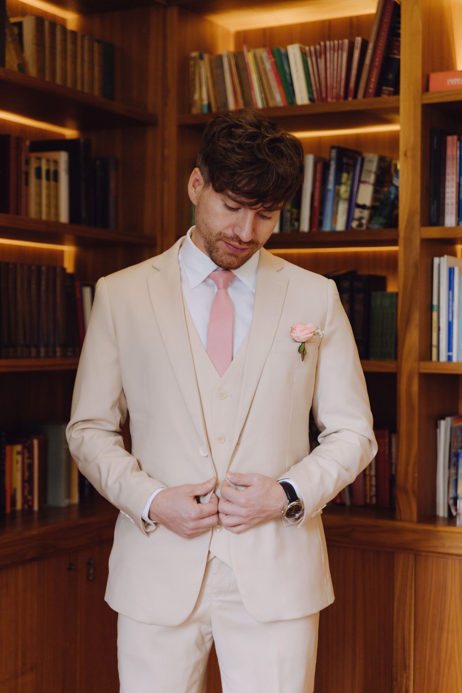Dapper groom in cream three-piece suit with pink tie and boutonniere buttoning jacket in a library.