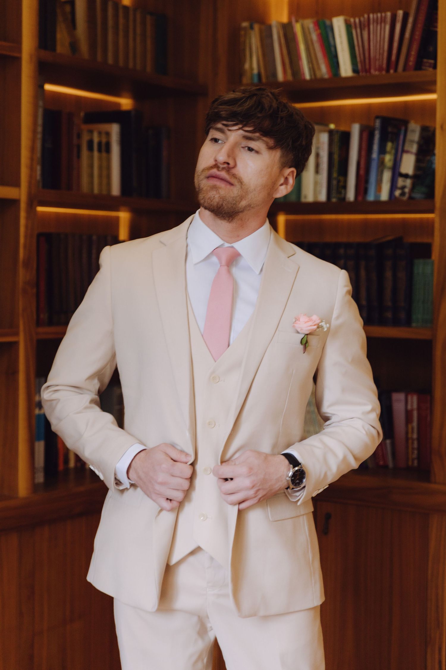 Confident groom in cream three-piece suit with pink tie and rose boutonniere posing in a library.