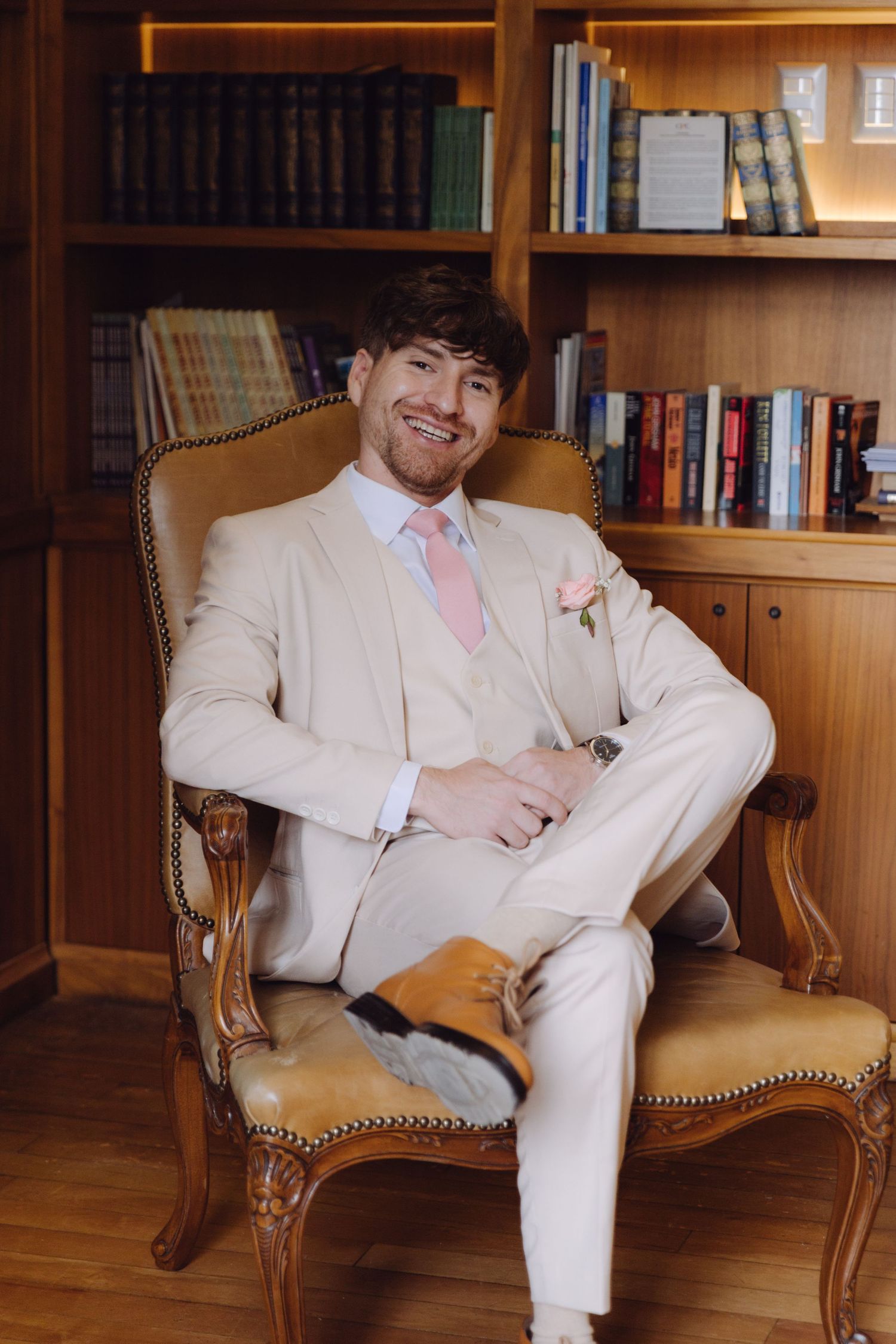Smiling groom in cream suit with pink tie sits relaxed in ornate leather chair in wood-paneled library.