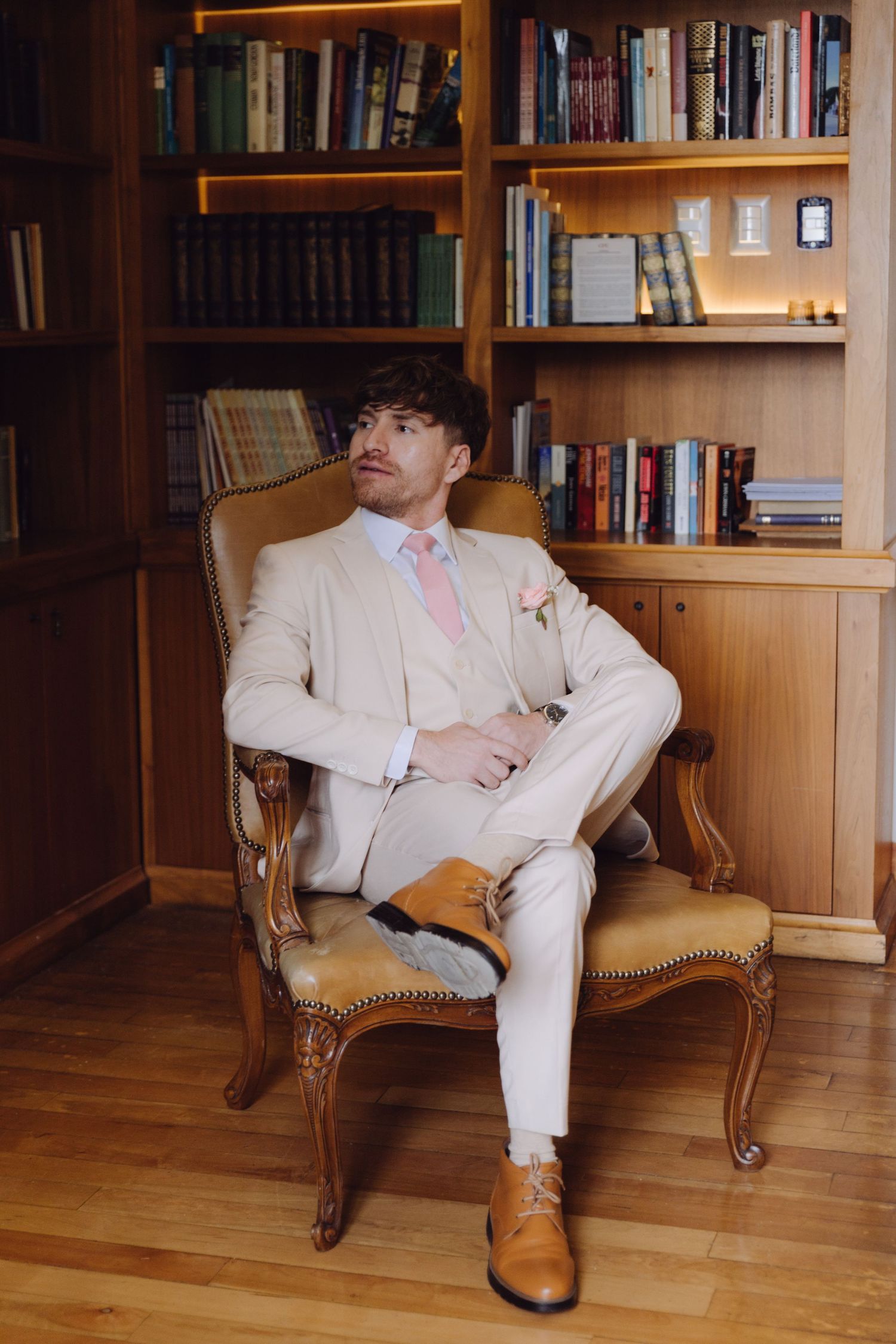 Stylish groom in cream three-piece suit with pink tie seated in vintage chair in a wood-paneled library.