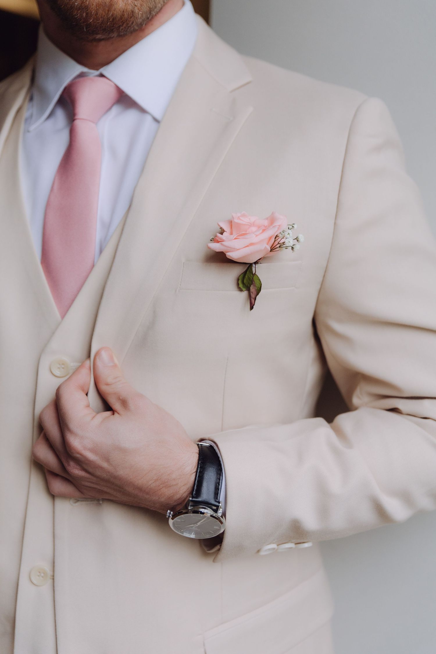 Groom in cream suit adjusting jacket with pink rose boutonniere and watch.