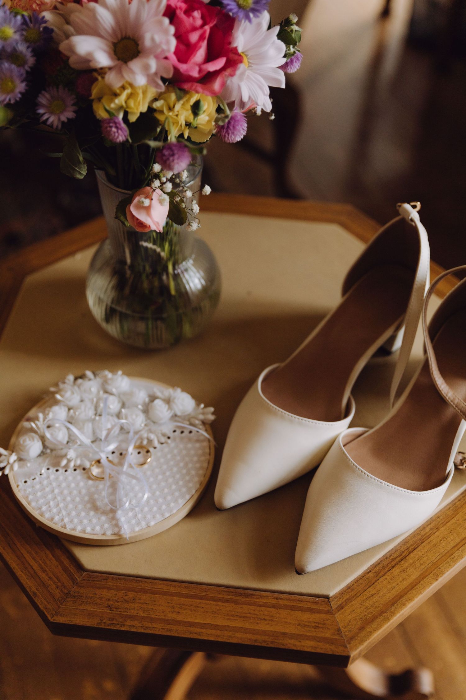 Bridal accessories with white heels, ring pillow, and colorful flower bouquet on wooden table.