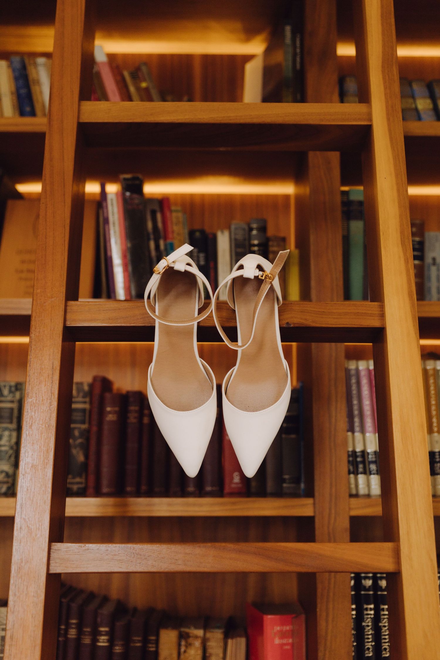 White bridal heels with ankle straps displayed on wooden bookshelf ladder.