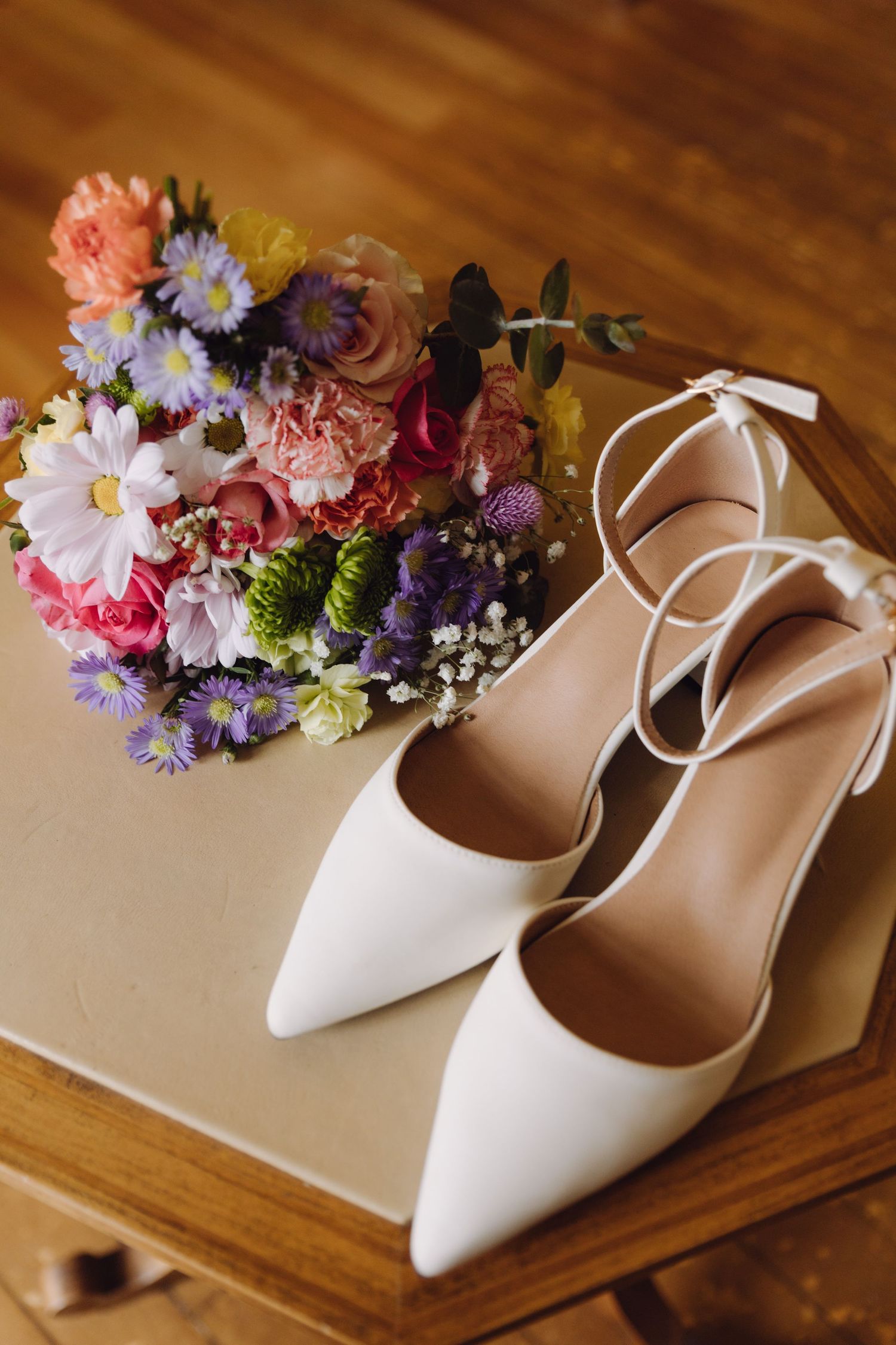 White pointed-toe bridal heels beside colorful wedding bouquet on wooden surface.