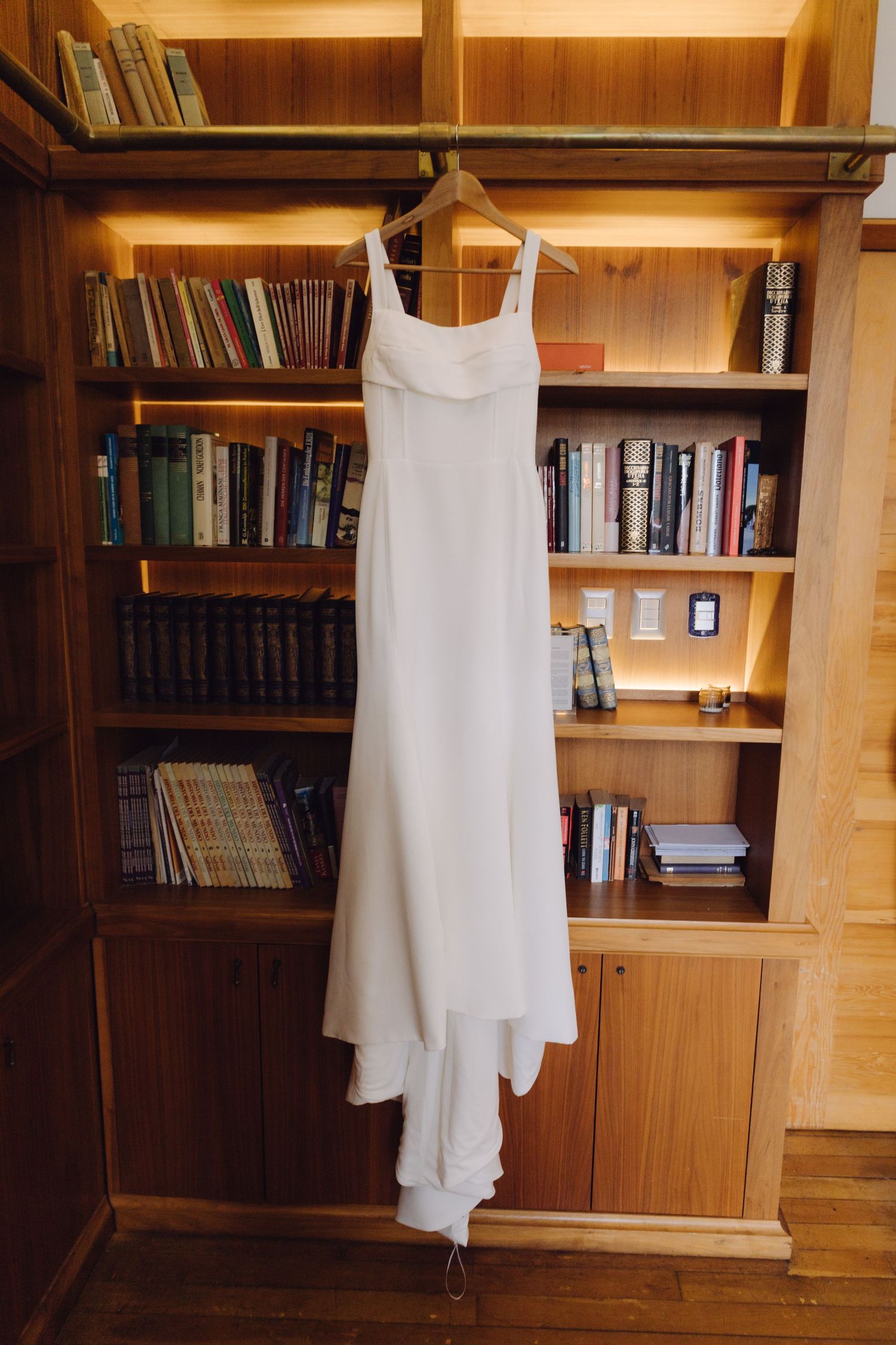 Elegant white wedding dress hanging on wooden bookshelf in warm lit room.