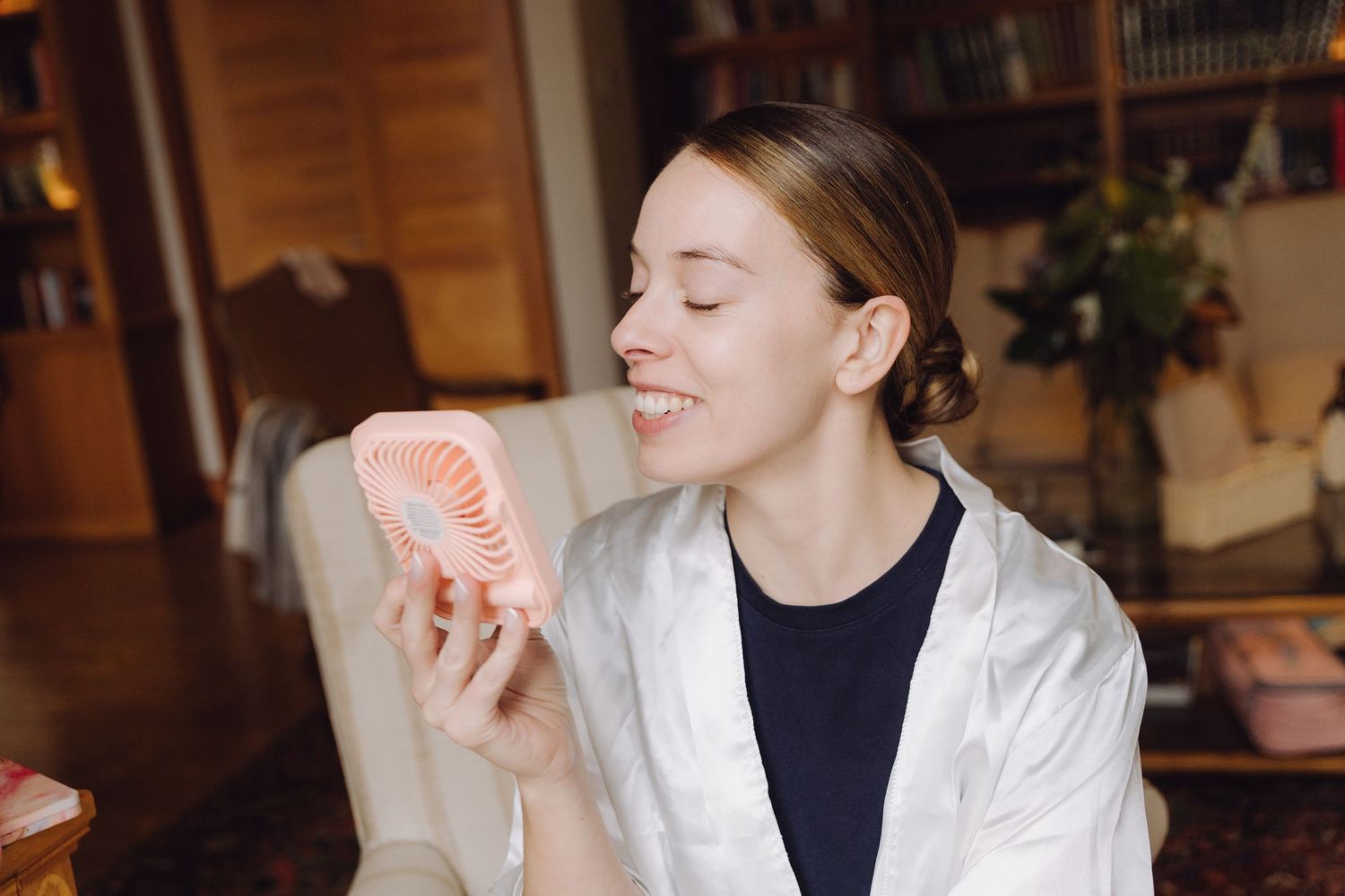 Smiling bride in white robe holding pink mini fan during wedding preparations.