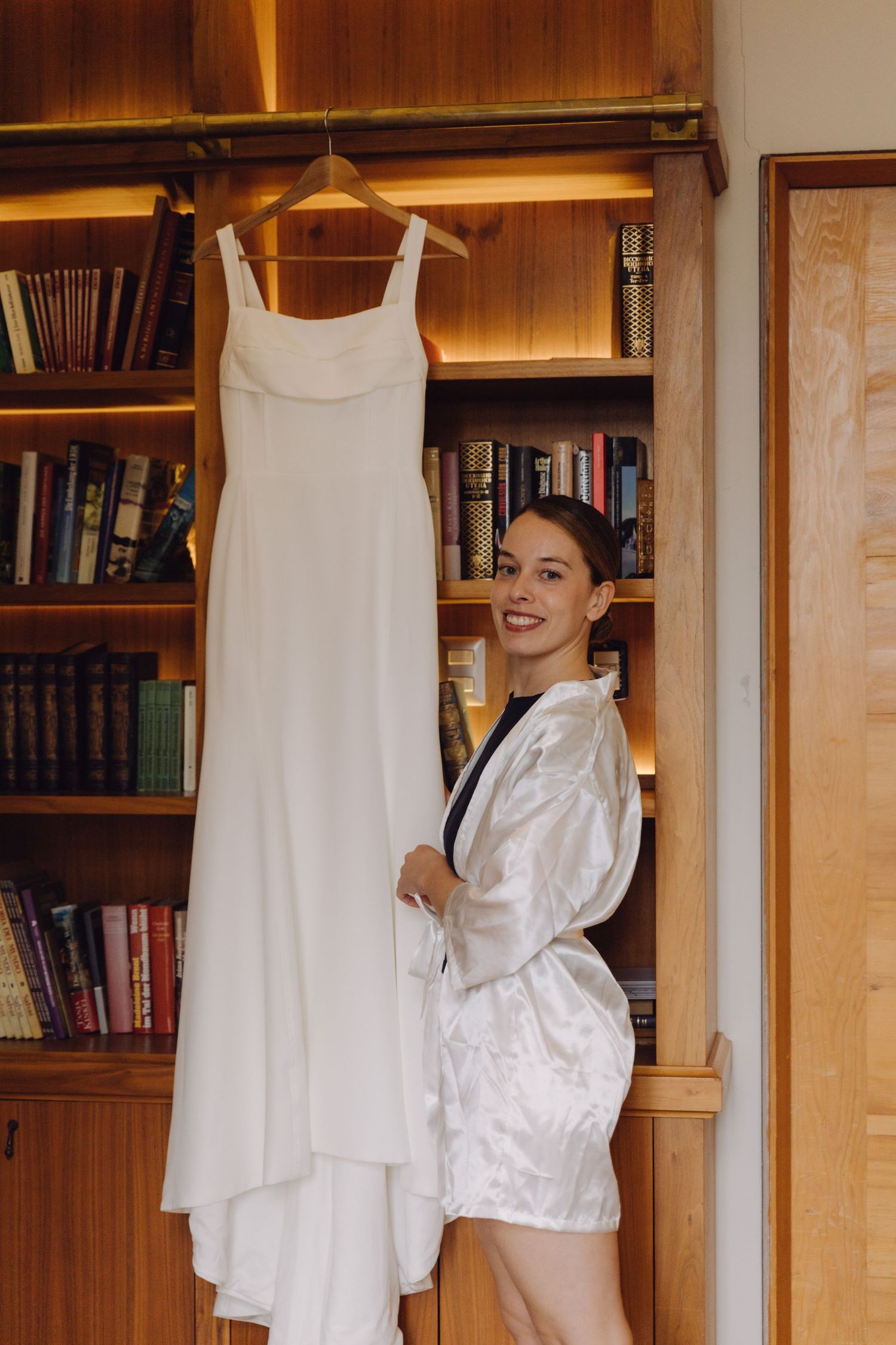 Happy bride in robe smiling beside hanging white wedding dress on bookshelf.