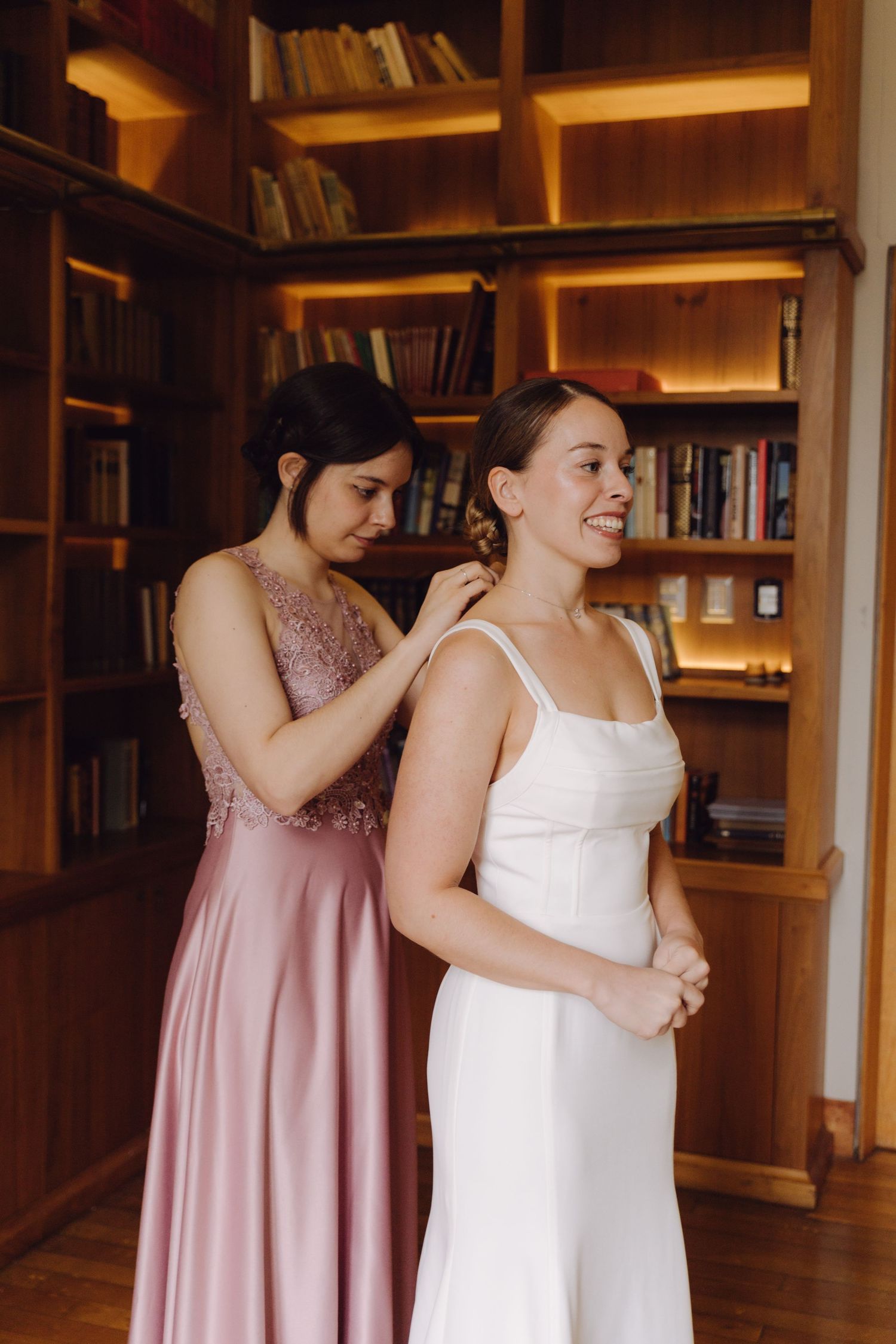Bridesmaid in pink dress helping bride with earring in front of bookshelf.