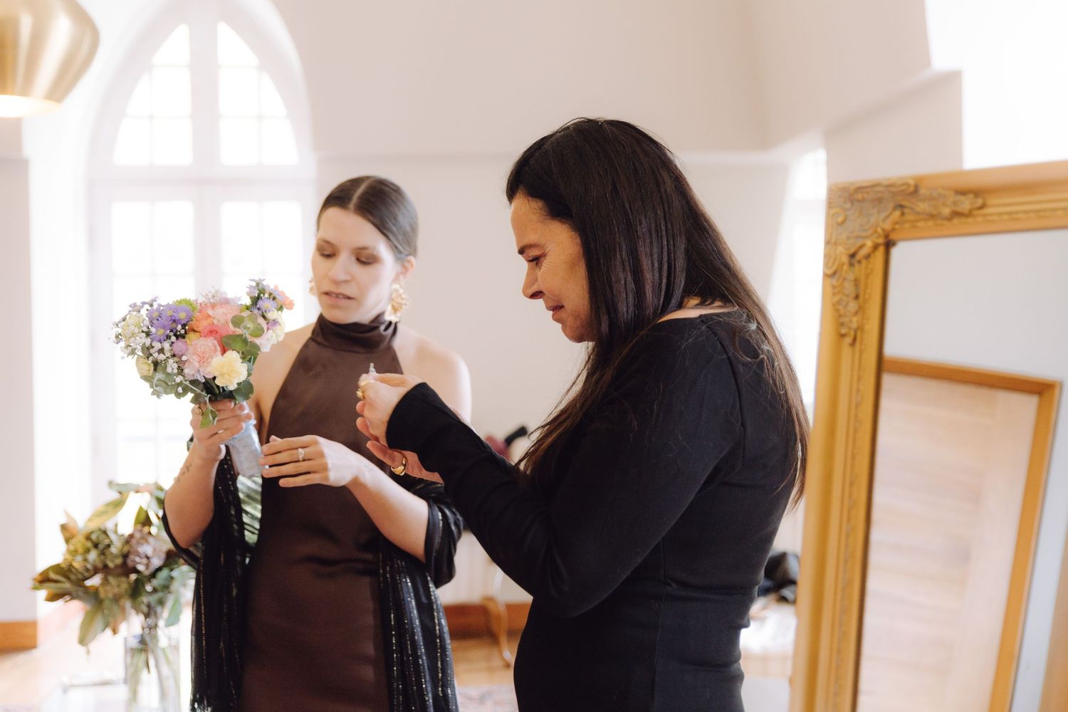 Wedding planner helping bride with dress details while holding a colorful bouquet in a bright room.