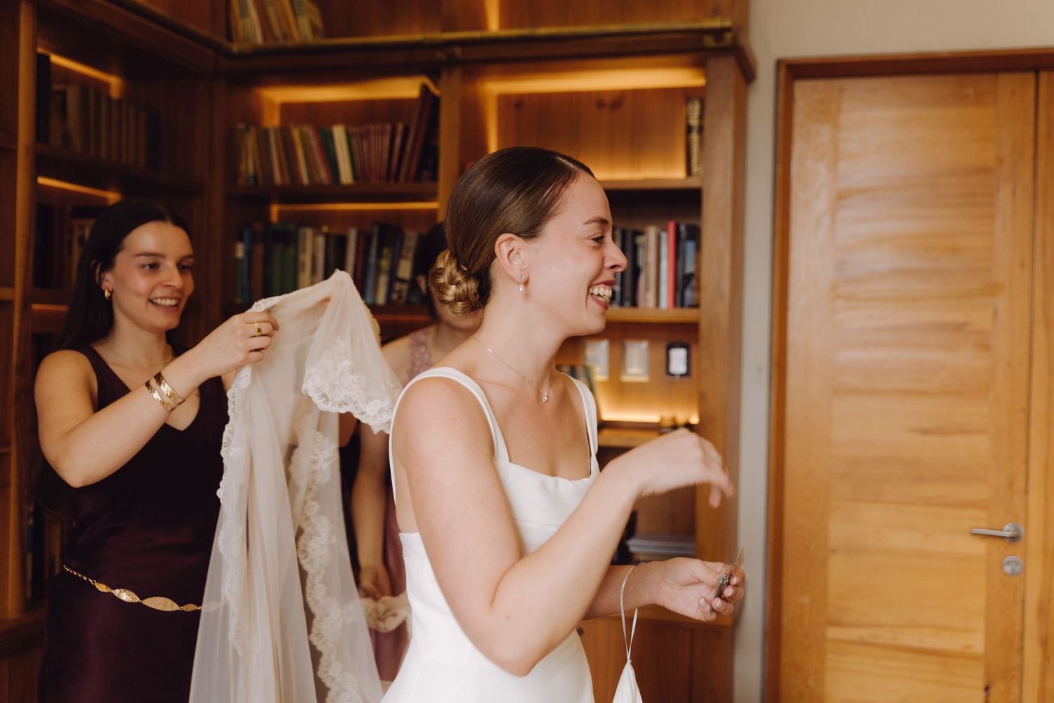 Bridesmaids laughing and helping smiling bride with her wedding veil in a wood-paneled library room.