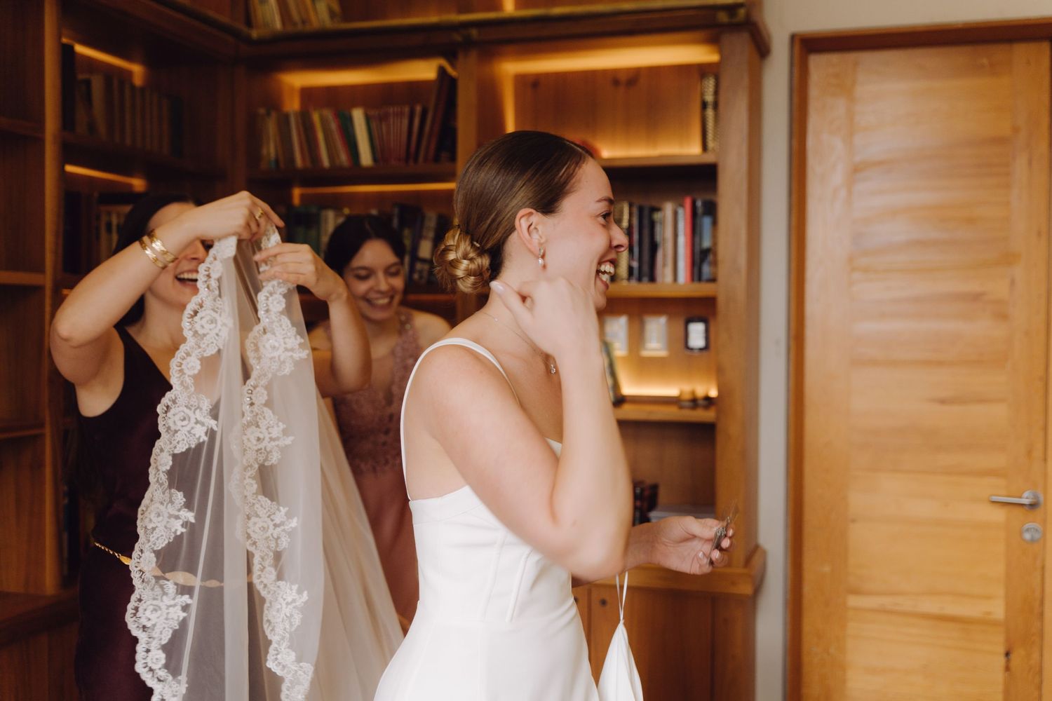 Bridesmaids holding lace-edged wedding veil behind smiling bride in a cozy library setting.