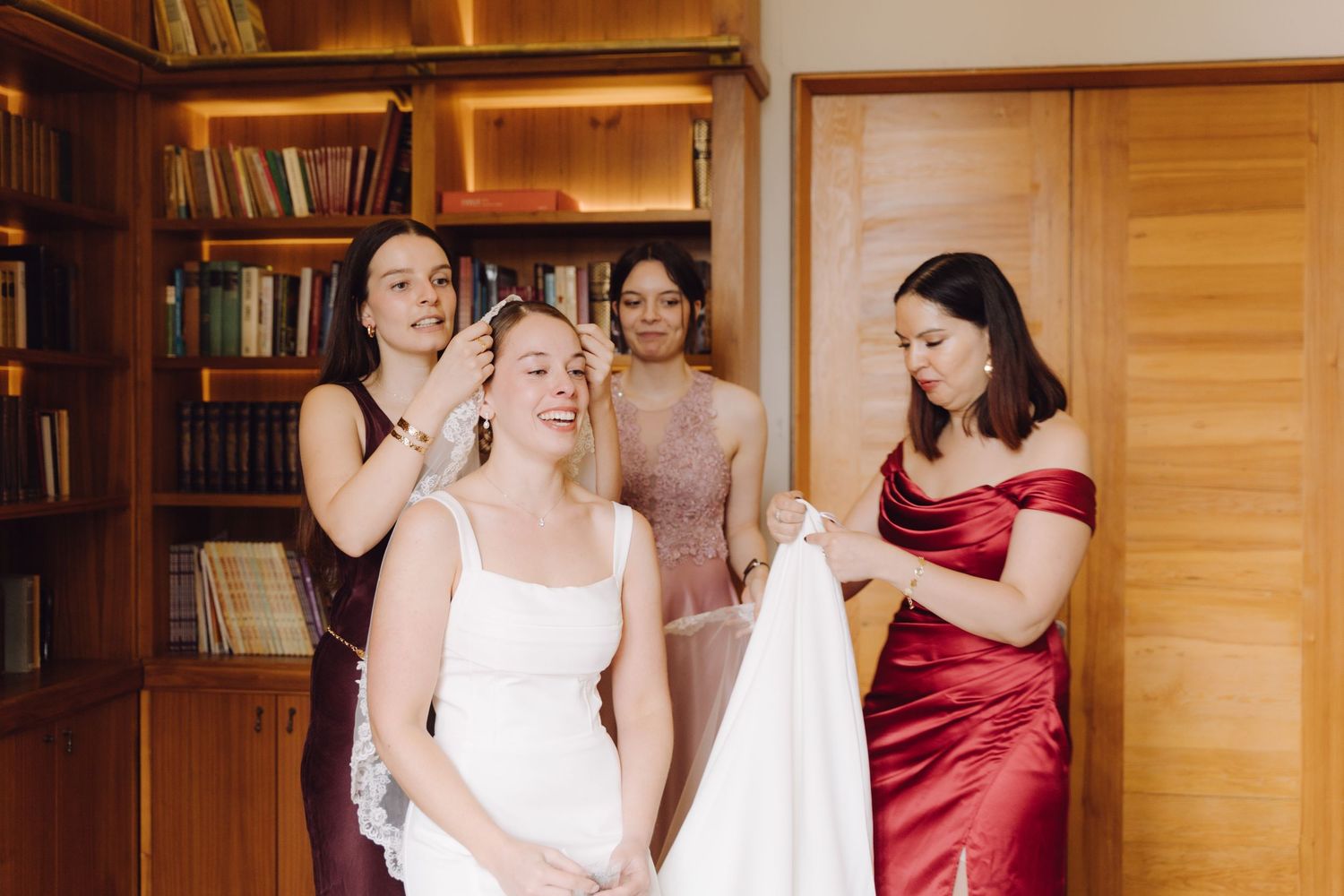 Three bridesmaids helping bride with hair and veil preparations in a warm library room.